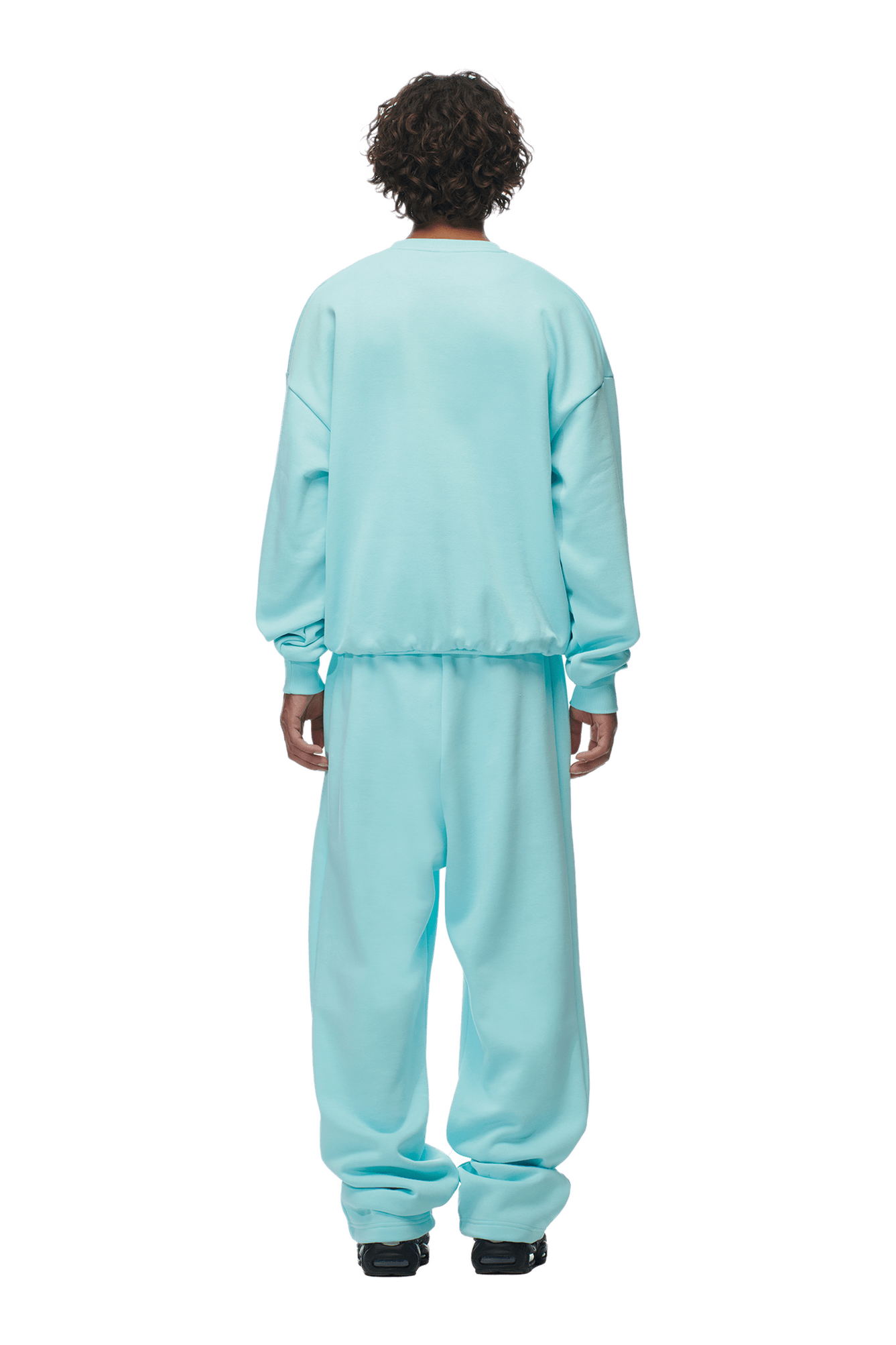 A person with curly hair faces away, wearing the 6PM CREWNECK TURQUOISE— a boxy fit sweatshirt with ribbed trims— and matching pants, paired with black shoes. The background is transparent.