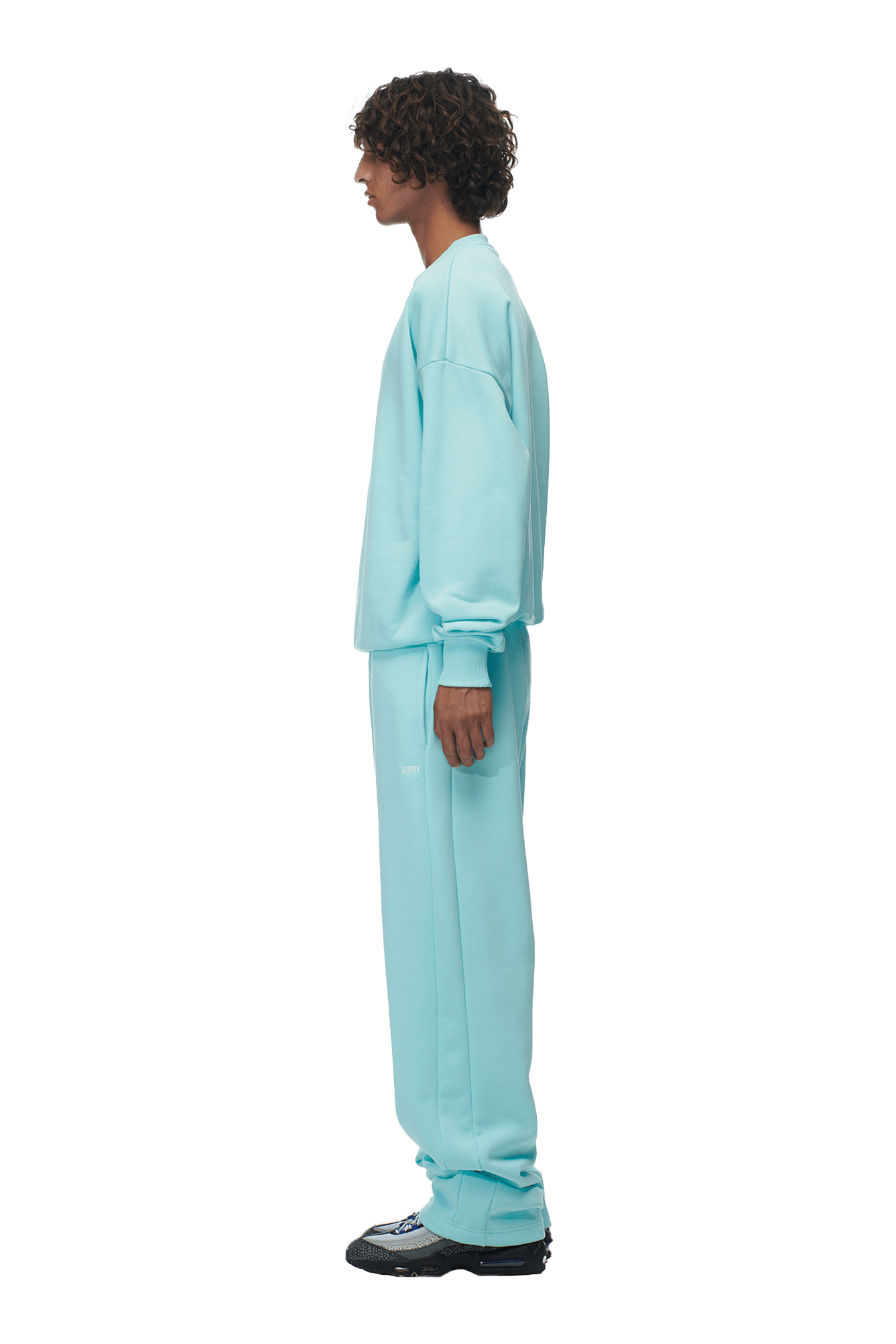 A person stands in profile facing left, wearing the 6PM CREWNECK TURQUOISE sweatshirt with ribbed trims and matching sweatpants, paired with black sneakers, against a plain background.