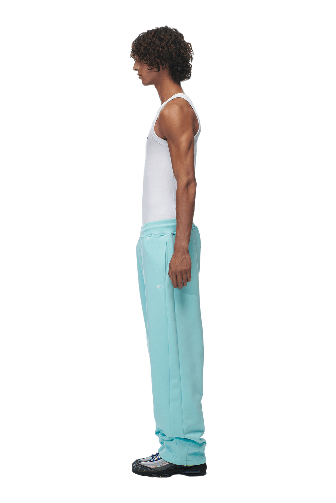 A man with curly hair stands in profile, wearing a white tank top, 6PMs OPEN LEG JOGGER TURQUOISE pants, and black shoes against a plain background.