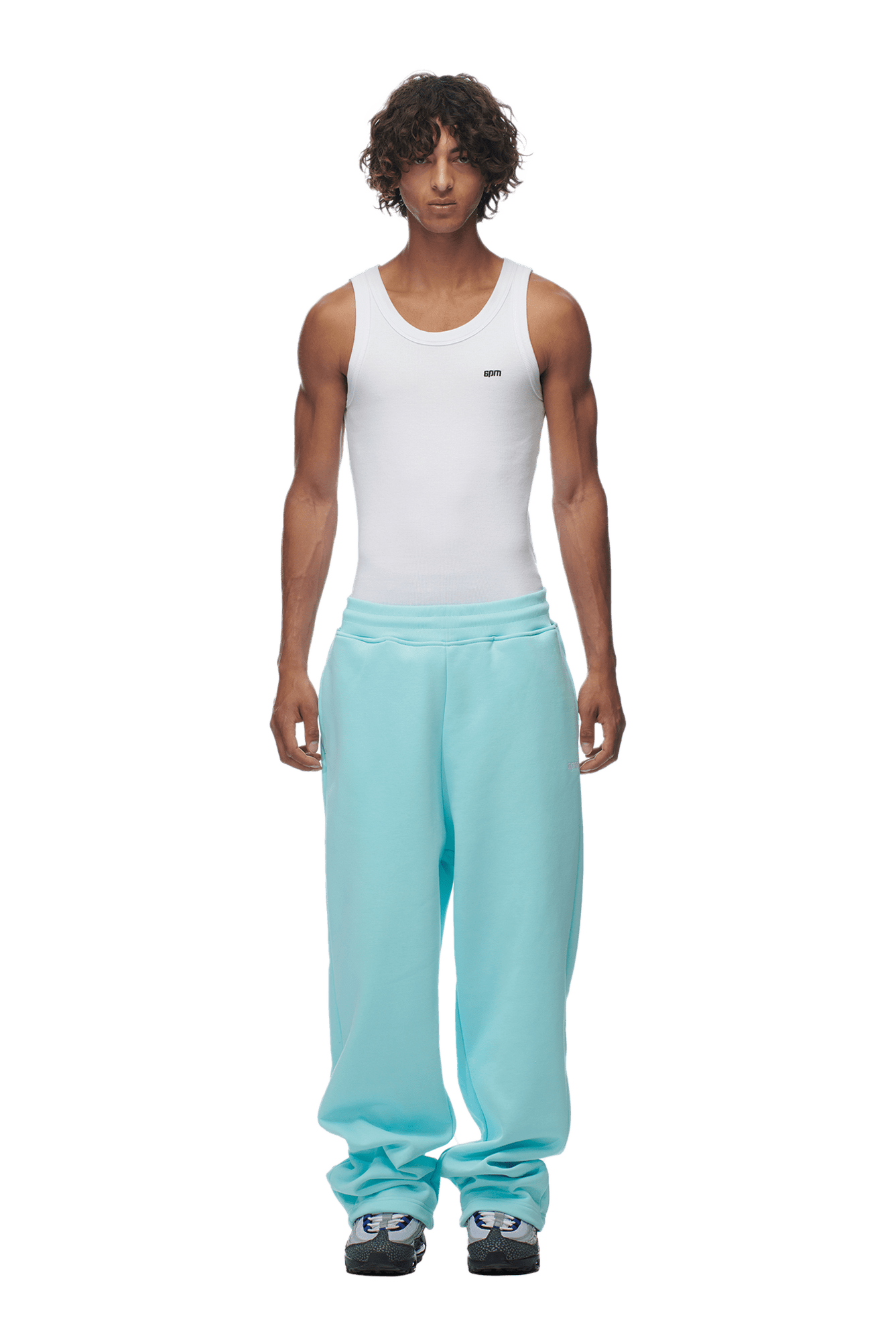 A person with curly hair wears a white tank top, 6PM OPEN LEG JOGGER TURQUOISE pants, and gray sneakers, standing against a plain background.