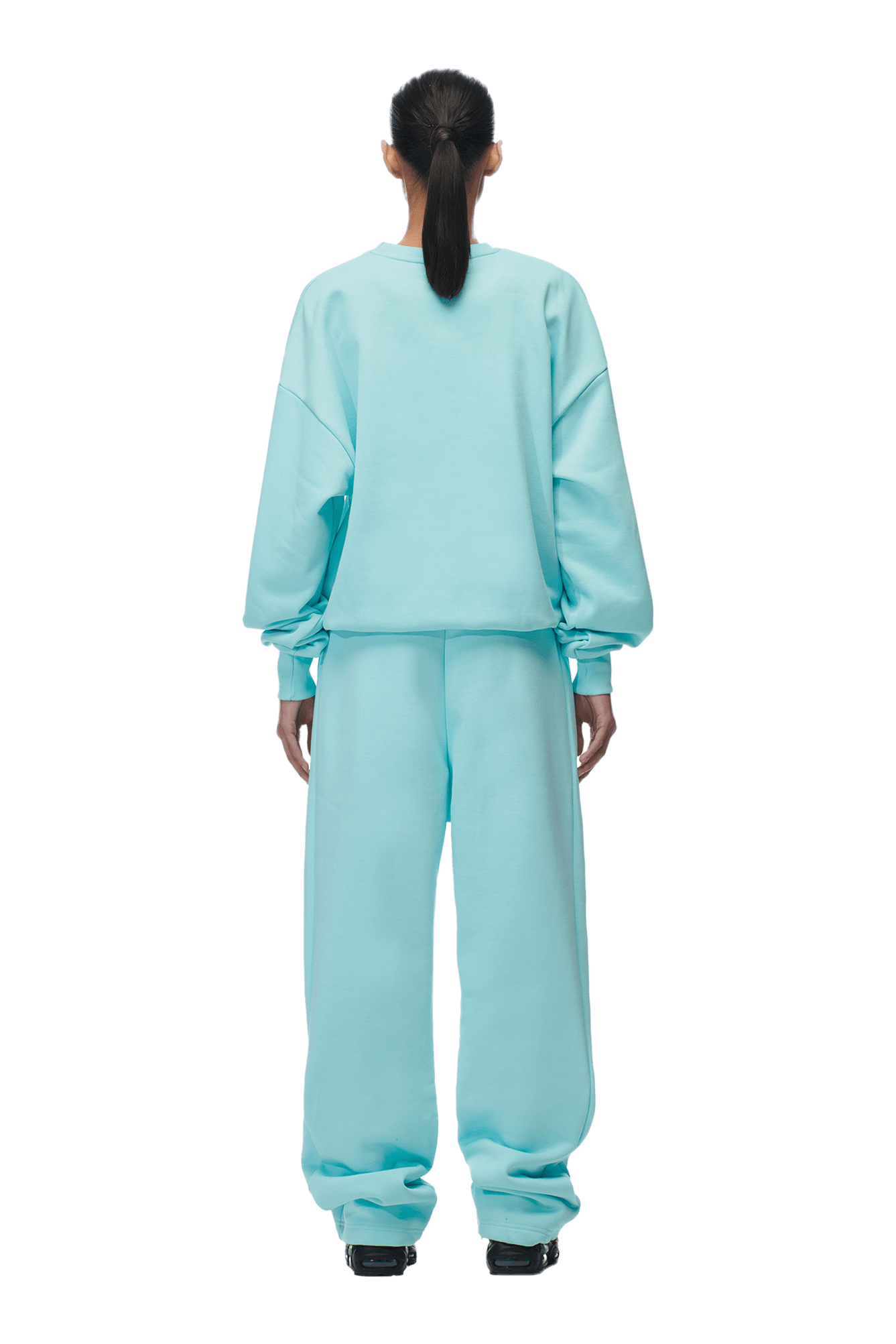 A person with a low ponytail stands with their back to the camera, wearing the 6PM CREWNECK TURQUOISE boxy fit sweatshirt and matching sweatpants, paired with black shoes. The image has a transparent background.
