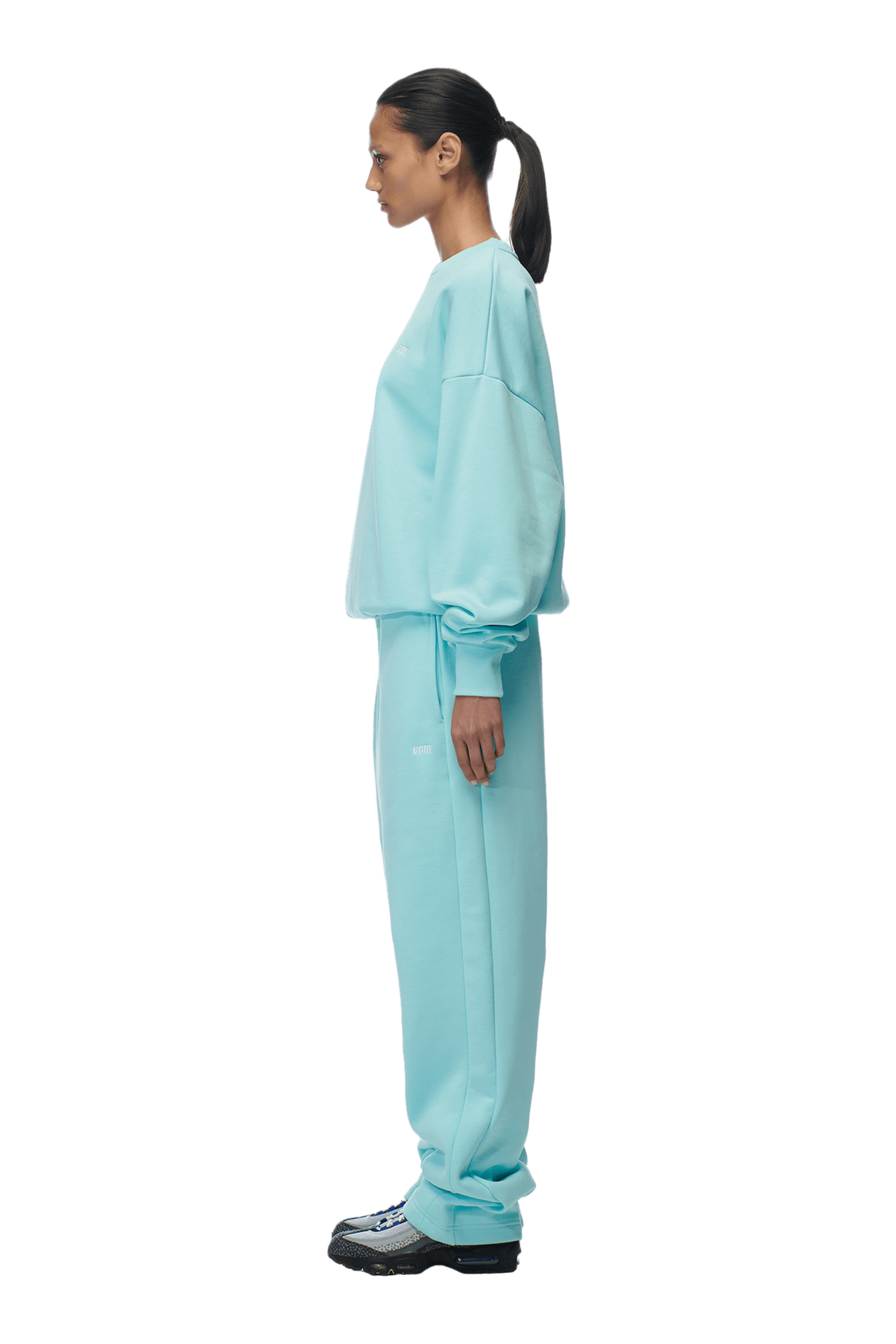 A woman with a ponytail stands in profile against a transparent background, wearing the 6PM CREWNECK TURQUOISE sweatshirt with matching sweatpants and black sneakers.