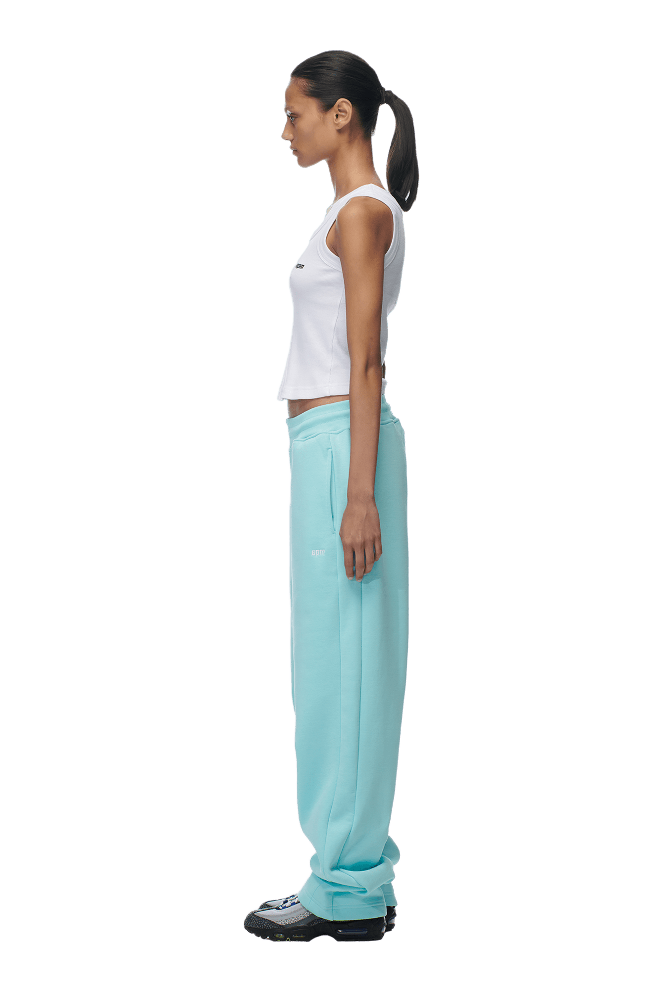 A woman in profile faces left, wearing a white sleeveless crop top, 6PM OPEN LEG JOGGER TURQUOISE pants, and black sneakers. Her dark hair is tied back in a low ponytail. The background is transparent.