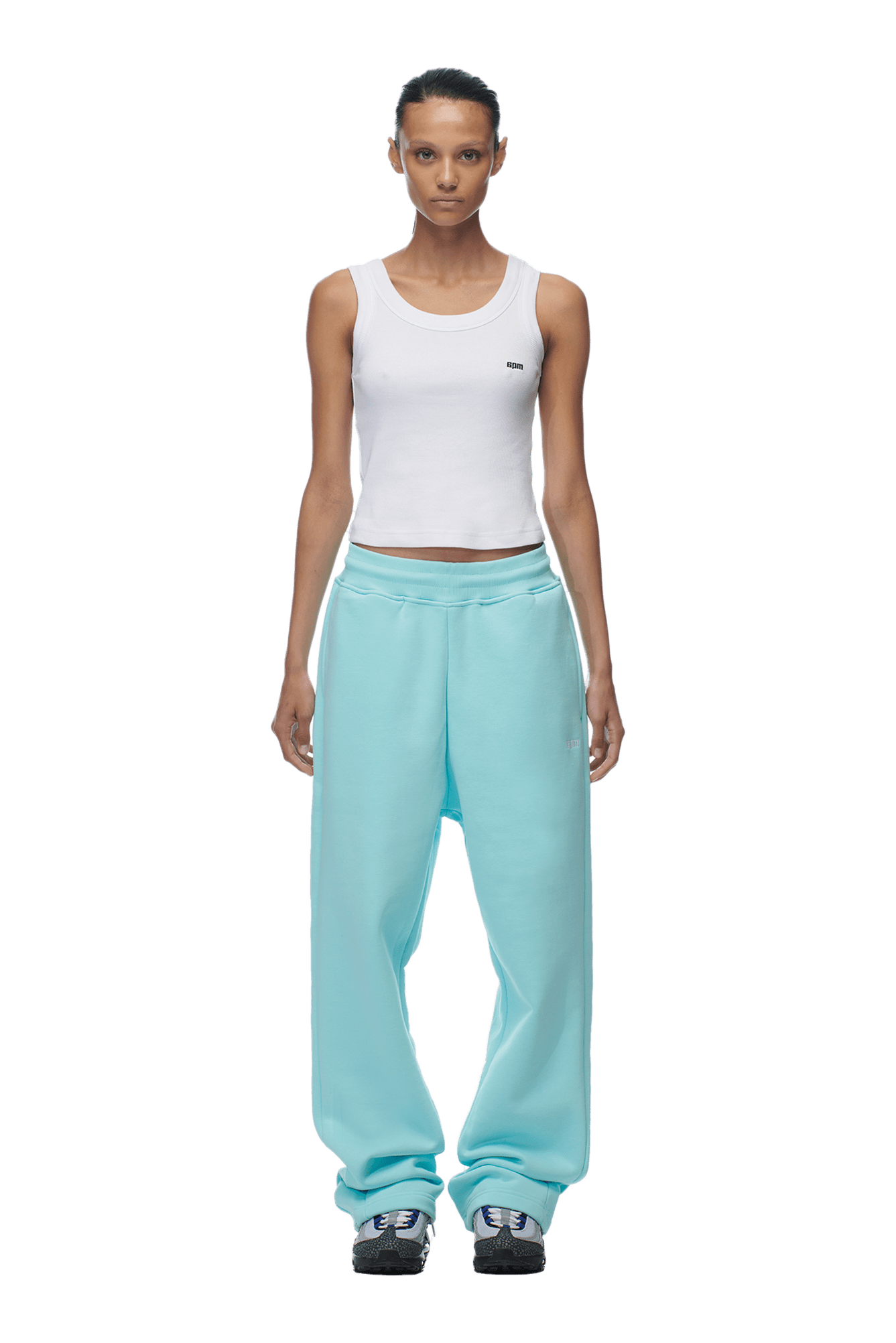 Against a plain green background, a person faces forward wearing a white sleeveless tank top, black and white sneakers, and 6PMs OPEN LEG JOGGER TURQUOISE pants.