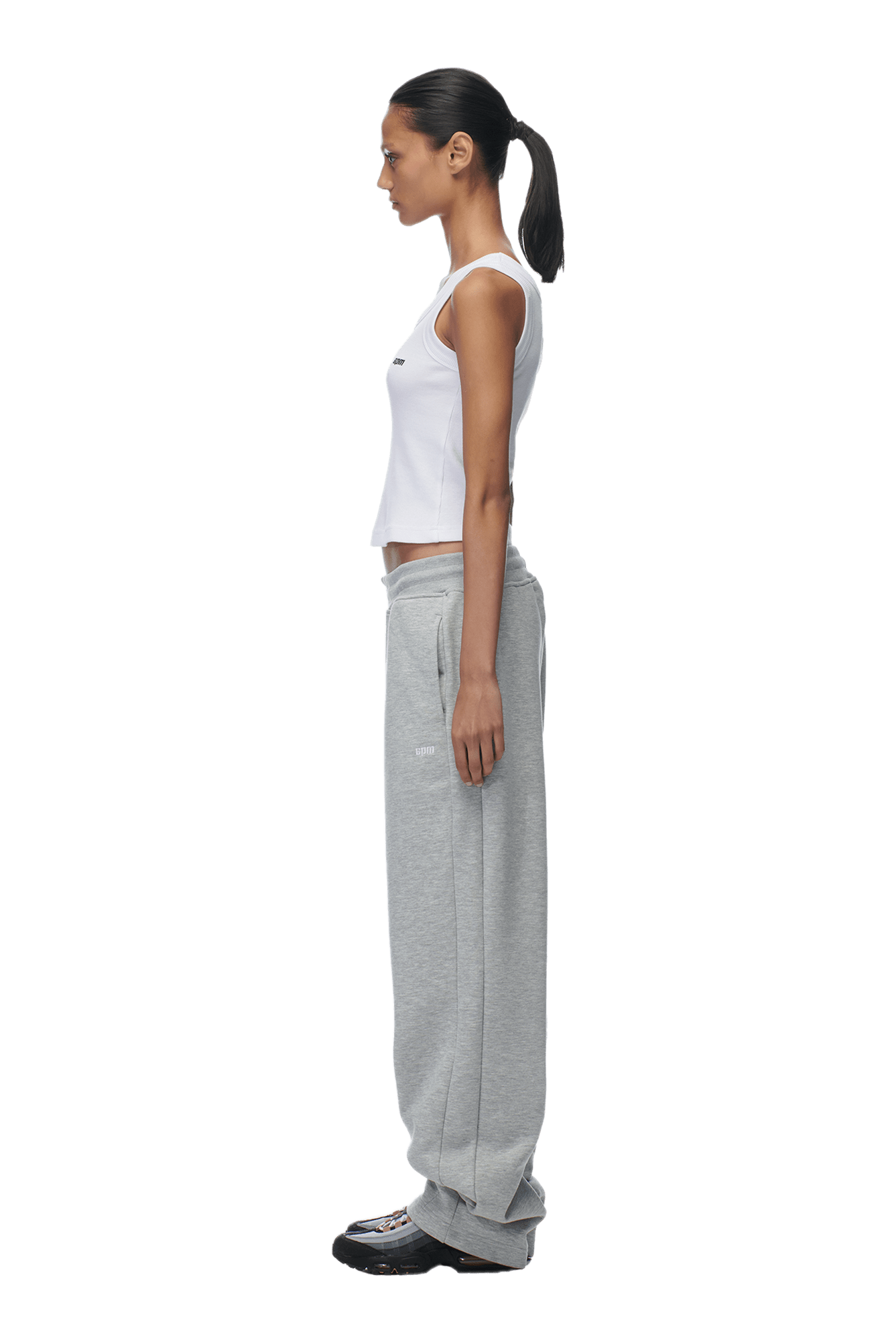 A woman in profile faces left, wearing a white tank top, 6PM OPEN LEG JOGGER GREY pants, and black sneakers. Her hair is tied back in a ponytail. The background is transparent.