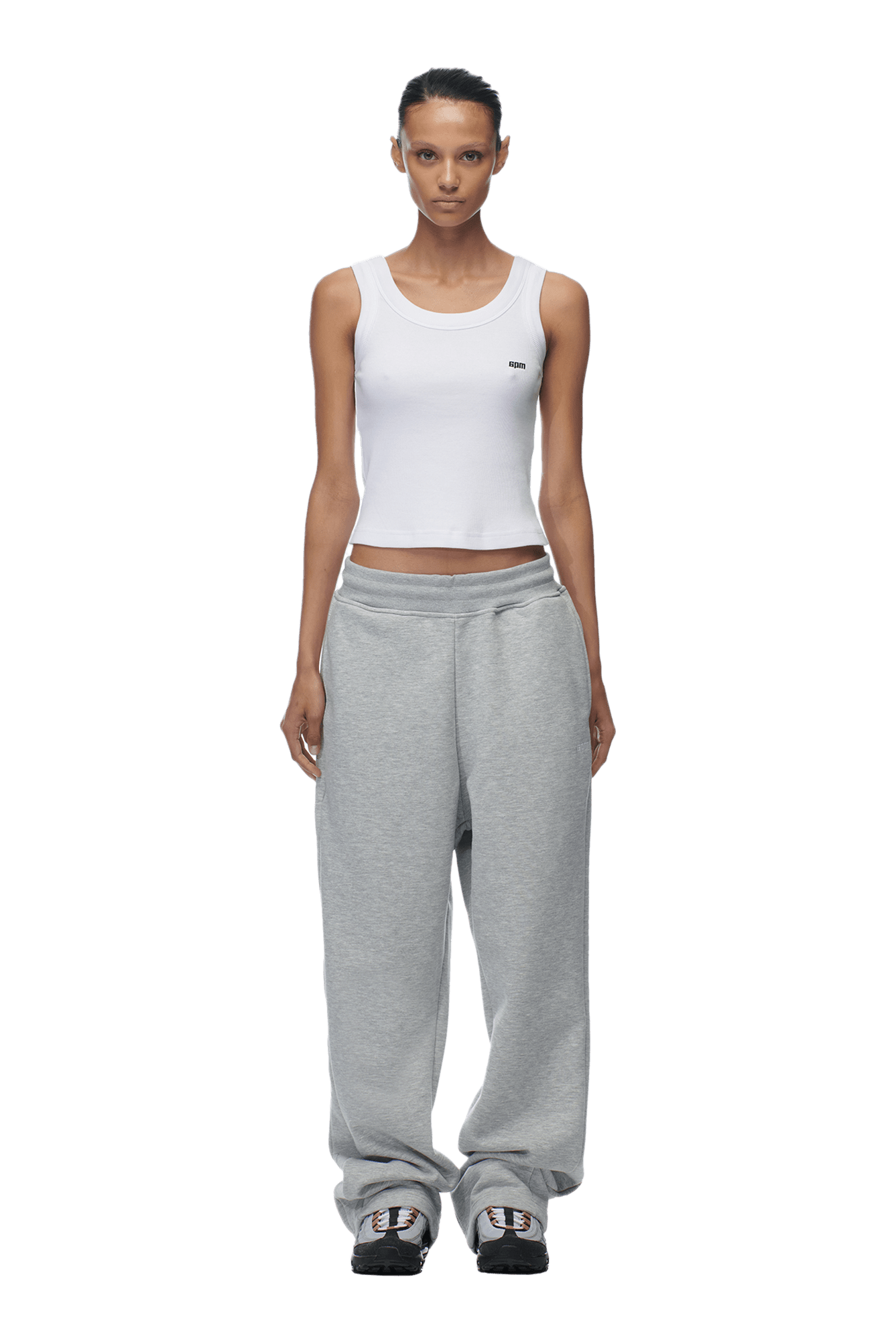 A woman with her hair pulled back and a neutral expression faces forward against a transparent background, wearing 6PMs OPEN LEG JOGGER GREY, a white sleeveless crop top, and gray sneakers.