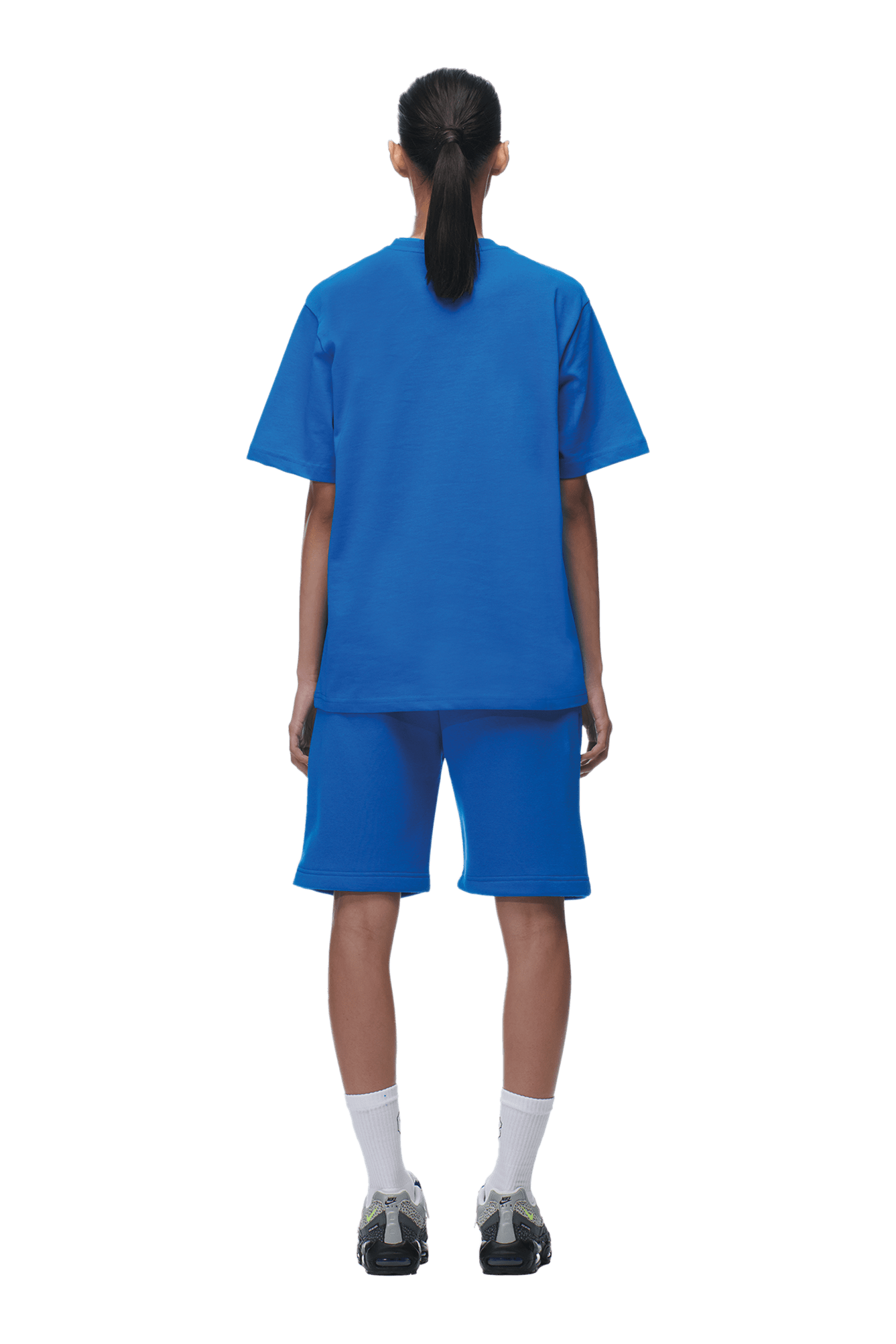 Individual with long dark hair in a ponytail, wearing the 6PM T-SHIRT PRINCESS BLUE, blue shorts, white socks, and dark sneakers, stands facing away from the camera on a transparent background—perfect for product showcases.