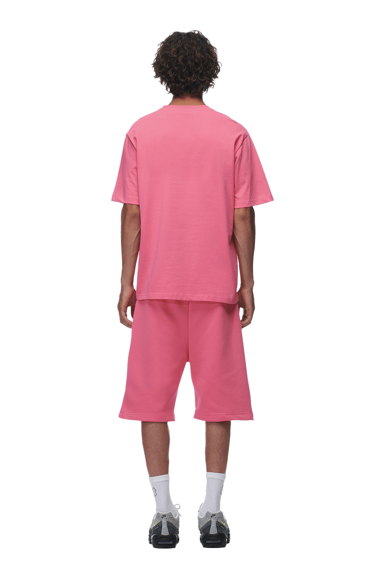 A person with curly hair faces away on a plain background, wearing the 6PM T-SHIRT HOT PINK with matching long shorts, white socks, and black sneakers.