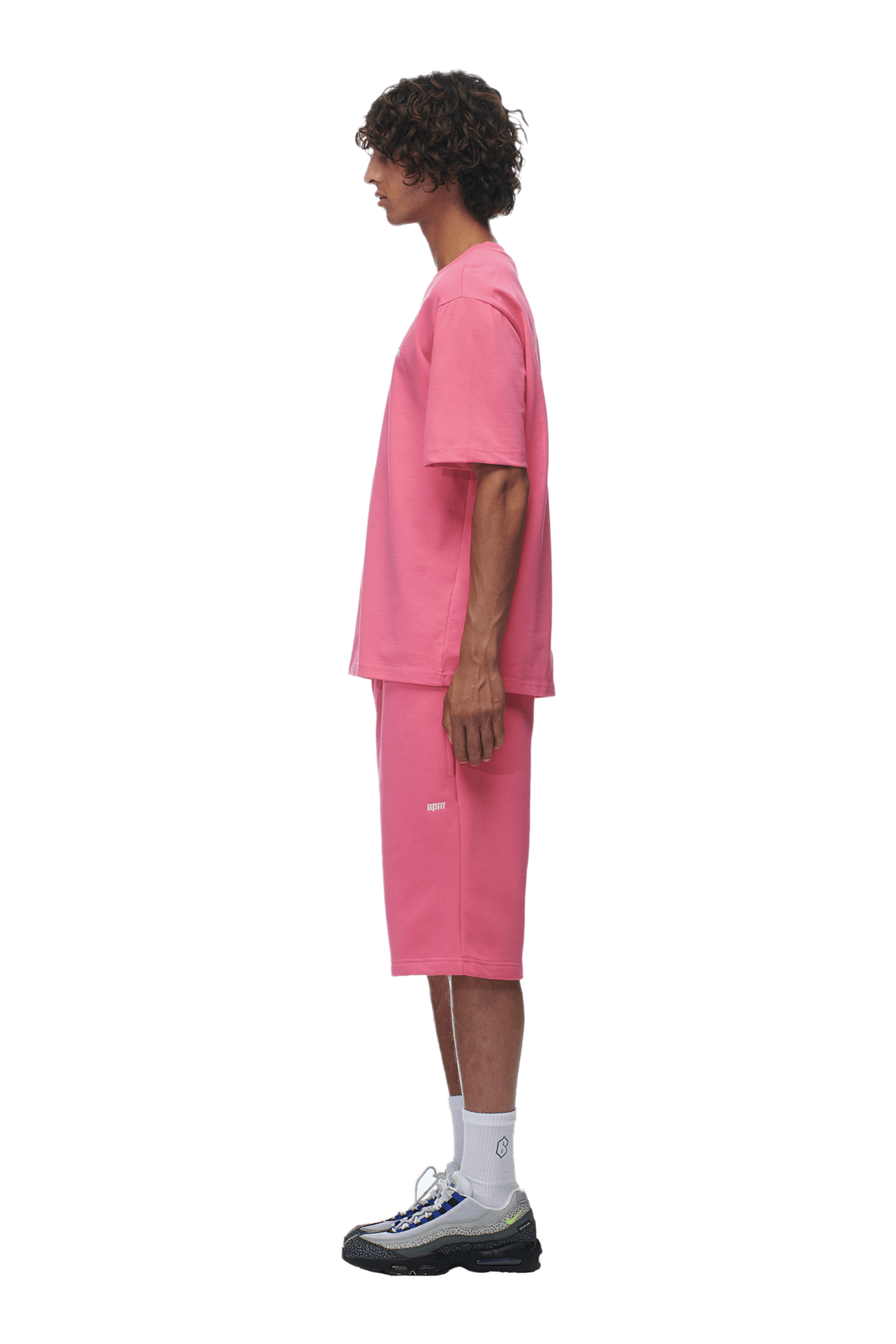 A person with curly hair stands in profile, wearing the 6PM T-SHIRT HOT PINK and matching pink shorts, white crew socks, and black sneakers against a plain background.