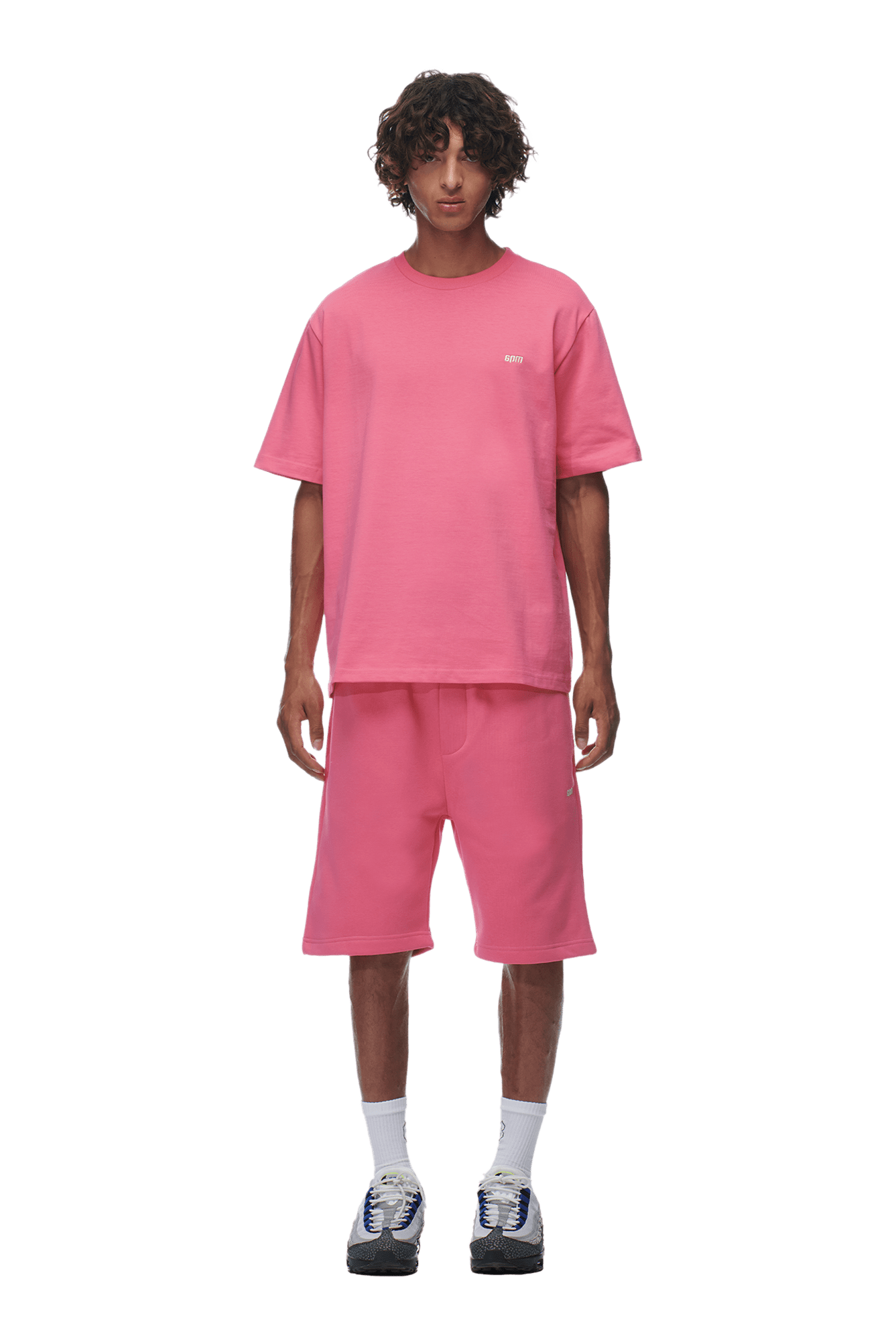 A person with curly hair stands facing forward, wearing a 6PM T-SHIRT HOT PINK and matching knee-length shorts, paired with white socks and sneakers, against a transparent background.