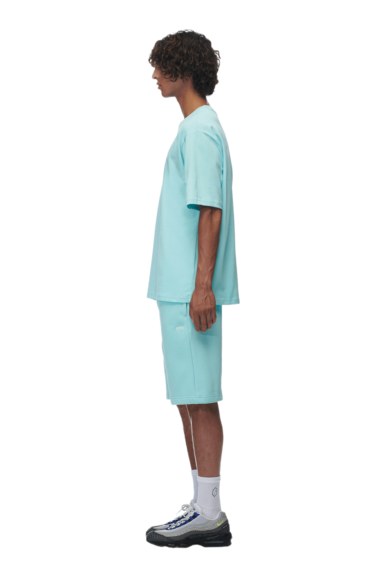 A young man with curly hair stands in profile, wearing the 6PM T-SHIRT TURQUOISE with matching shorts, white socks, and black sneakers against a plain background.