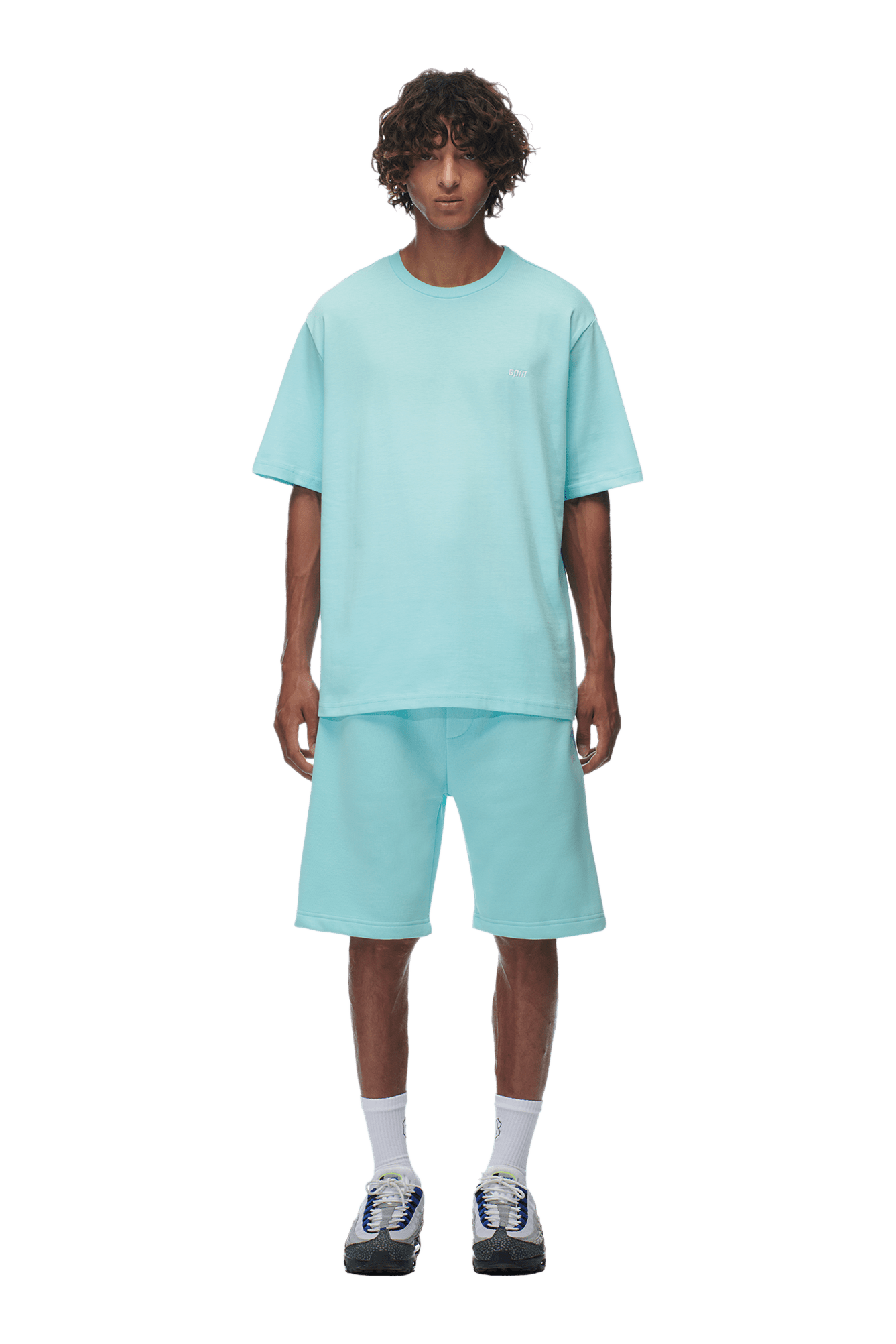 A person with curly hair wears the 6PM T-SHIRT TURQUOISE and matching shorts, with white socks and black-and-white sneakers, standing against a transparent background.