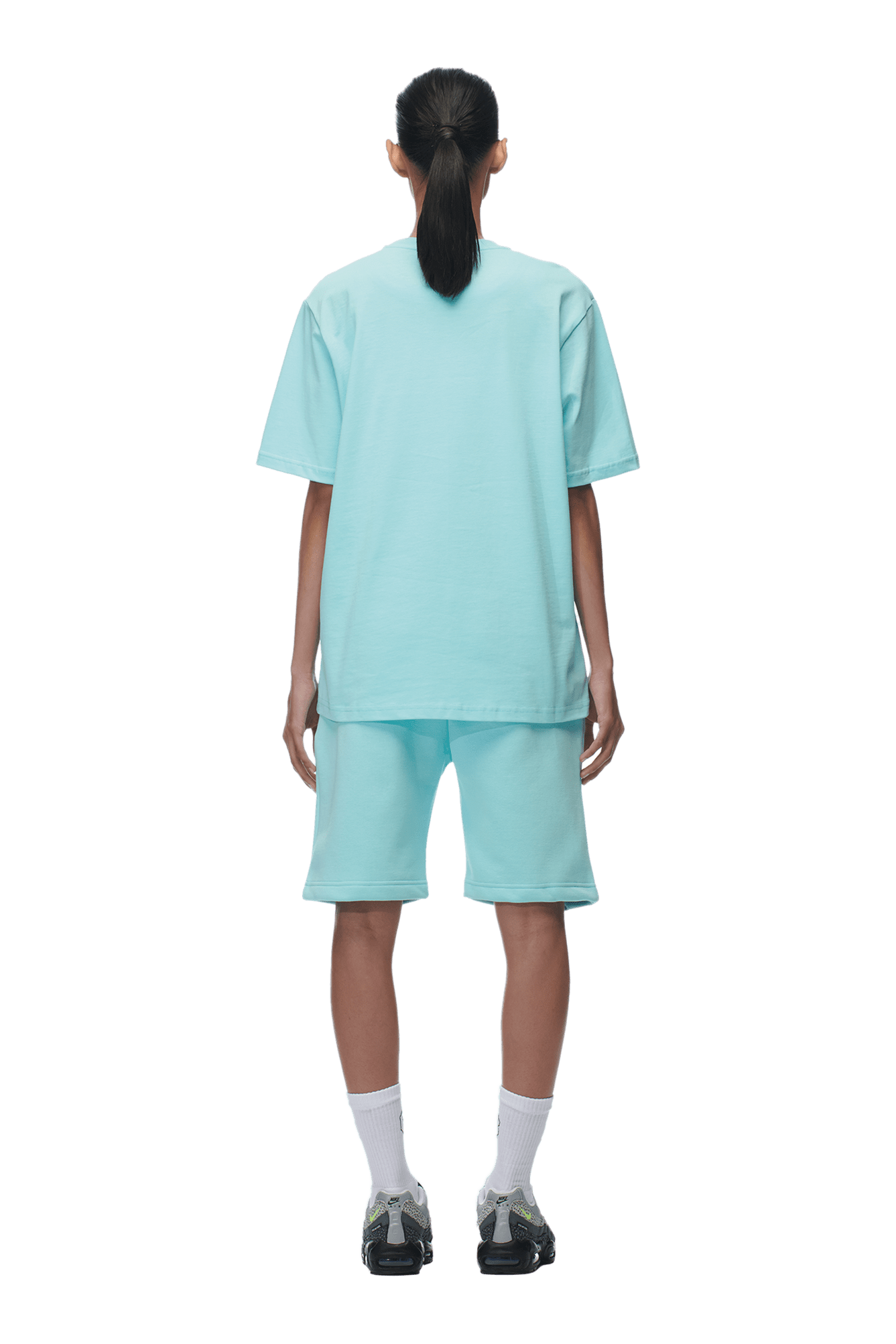A person with dark hair in a low ponytail stands facing away, wearing a loose-fitting 6PM T-SHIRT TURQUOISE and matching shorts, white socks, and black sneakers. The background is plain and transparent.