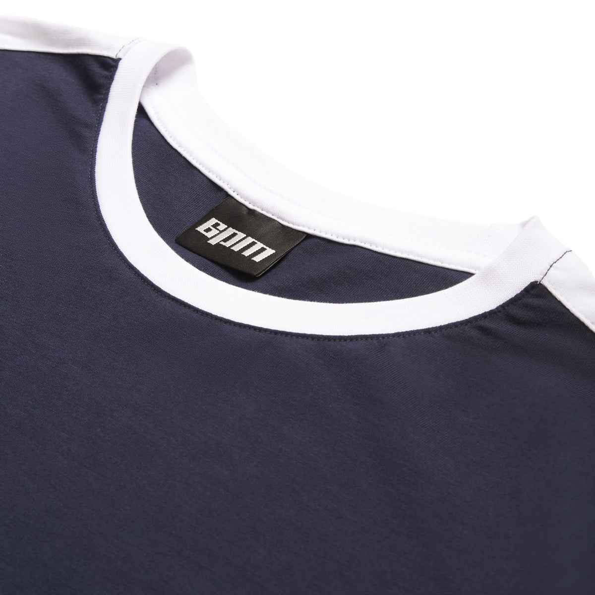 A close-up of the 6PM GRAFFITI RINGER LS MIDNIGHT BLUE shows its navy fabric, white collar, and a black label with white text sewn inside the neckline, laid flat on a white background.
