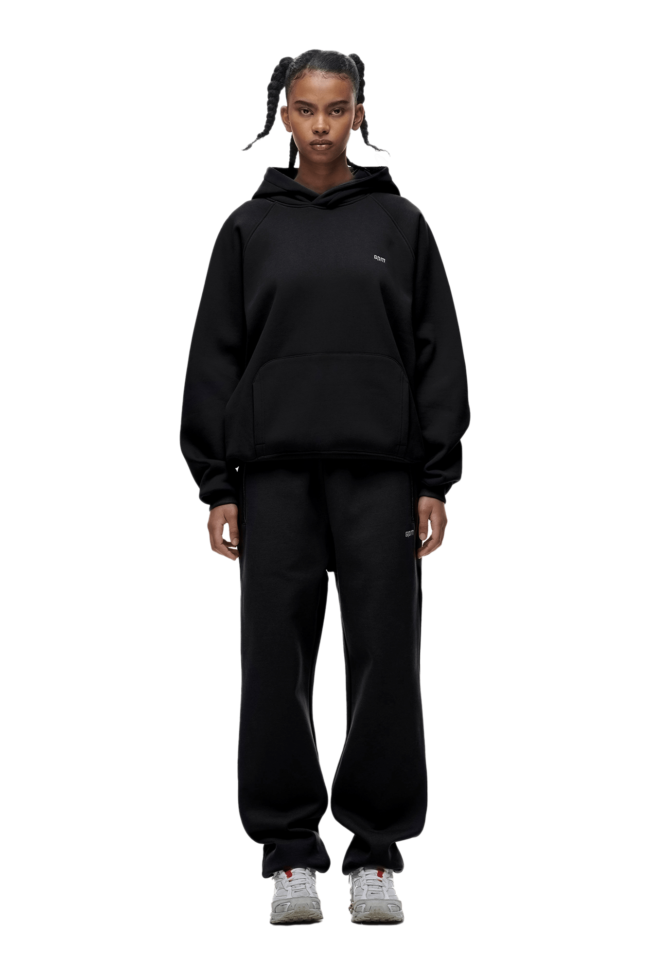 A person with braided hair faces forward, wearing the HOODIE BLACK—a loose black raglan-sleeved hoodie with a kangaroo pocket—plus matching black sweatpants and white sneakers. They stand against a transparent background, perfect for daily wear.