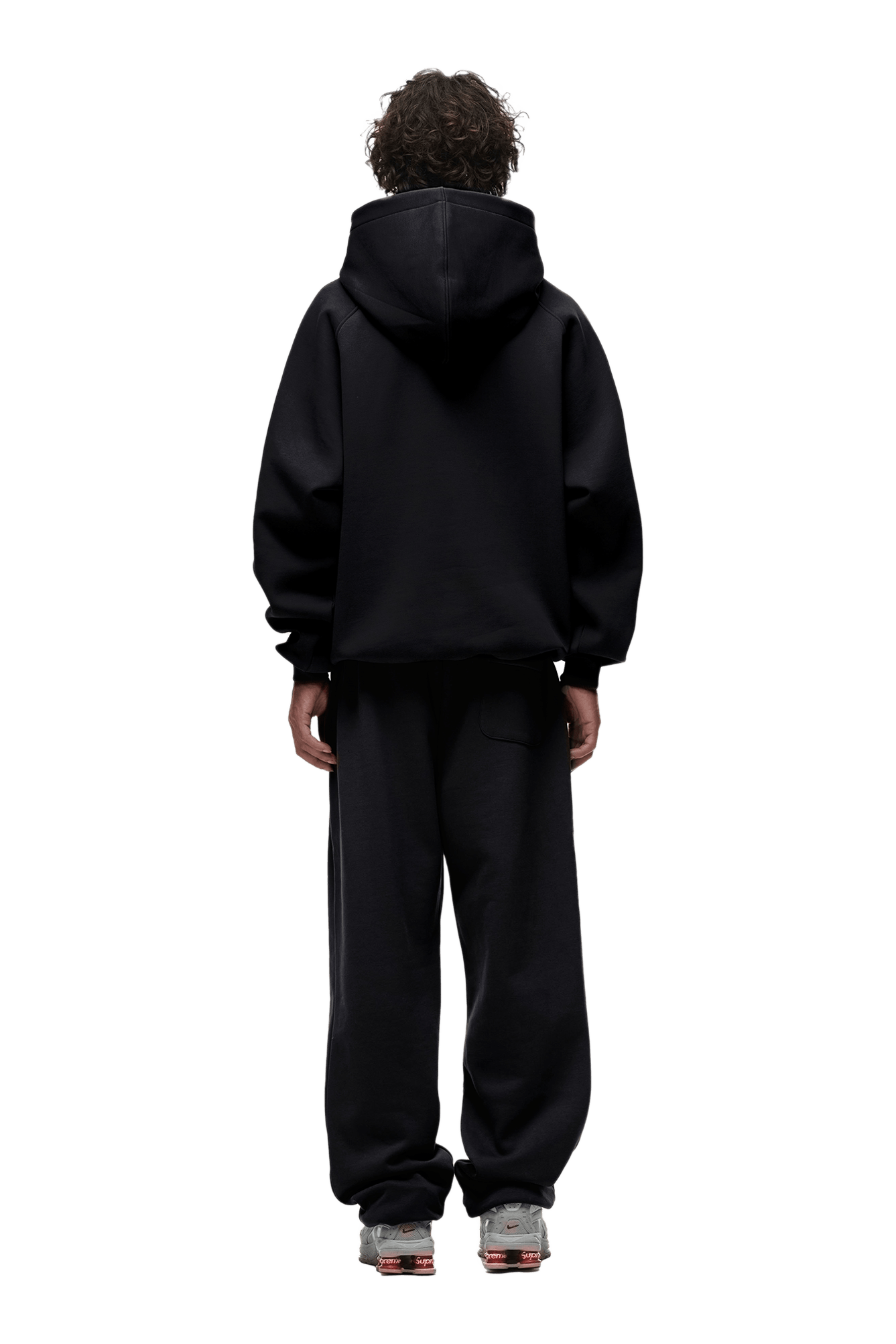 A person with curly hair stands facing away, wearing the HOODIE BLACK—featuring raglan sleeves and a kangaroo pocket—with black sweatpants and gray sneakers on a transparent background. Perfect for everyday wear.