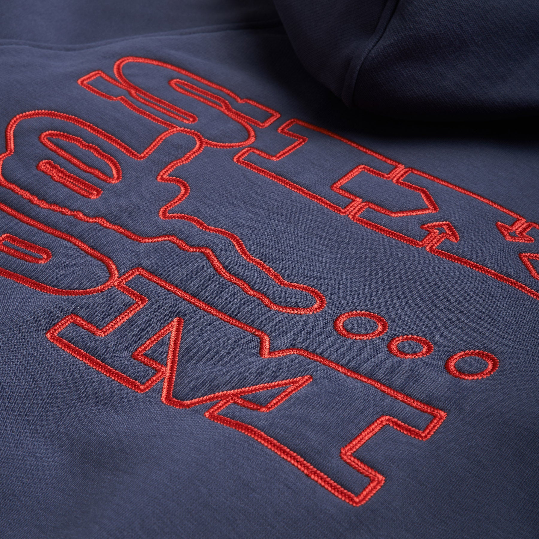 Close-up of the SLAWN ZIP HOODIE by 6PM, featuring navy blue fabric with bold red embroidered outline text and shapes. The raised stitching adds a textured effect, highlighting the details on this stylish hoodie.