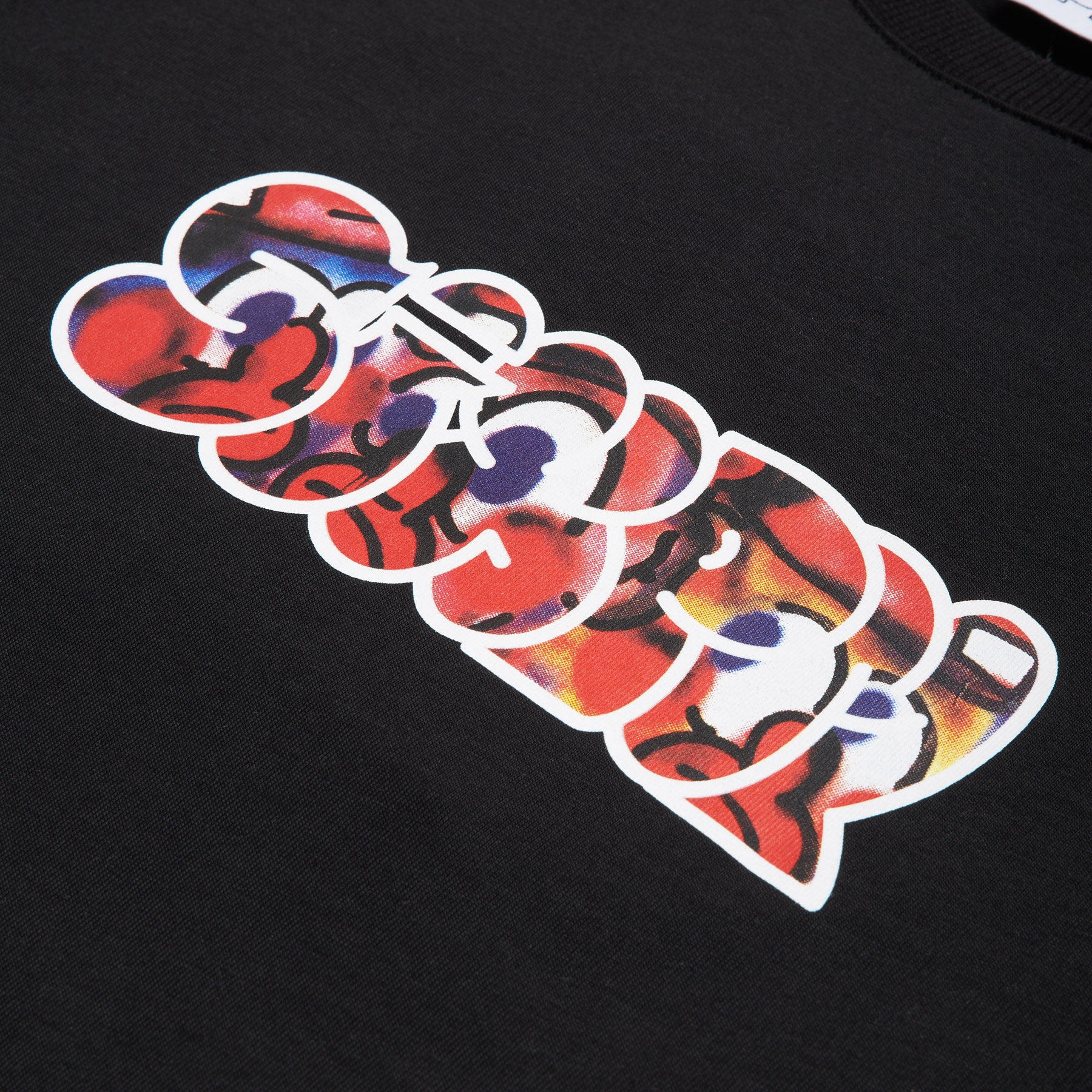 A close-up of the SLAWN SIGNATURE TEE BLACK by 6PM, showcasing its black fabric with a vibrant, abstract graffiti-style print in red, yellow, purple, and black, highlighted by bold white outlines.