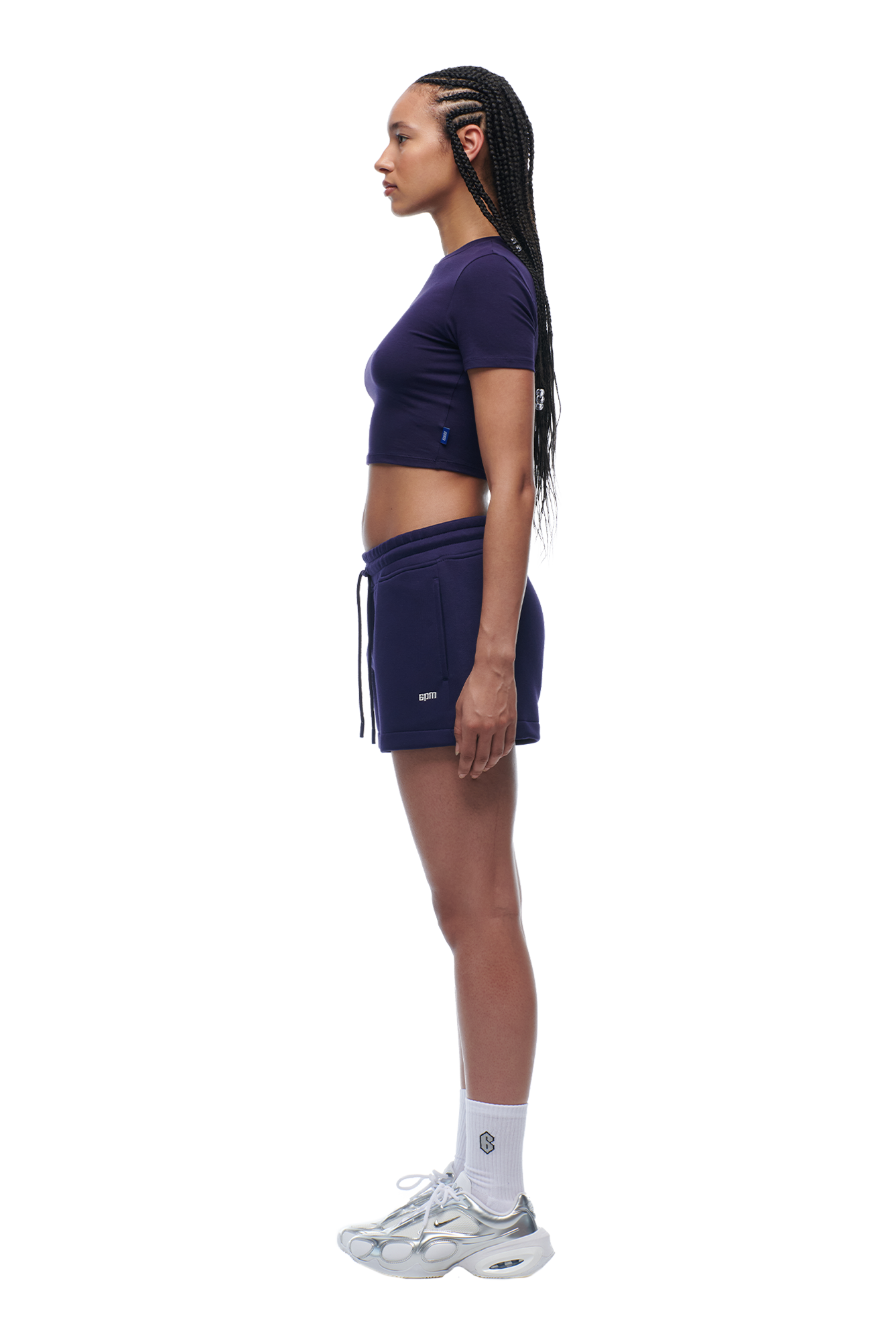 A woman with braided hair stands in profile wearing the 6PM WMNS CROPPED T-SHIRT ORBIT BLUE, matching shorts, white socks, and silver sneakers against a white background.