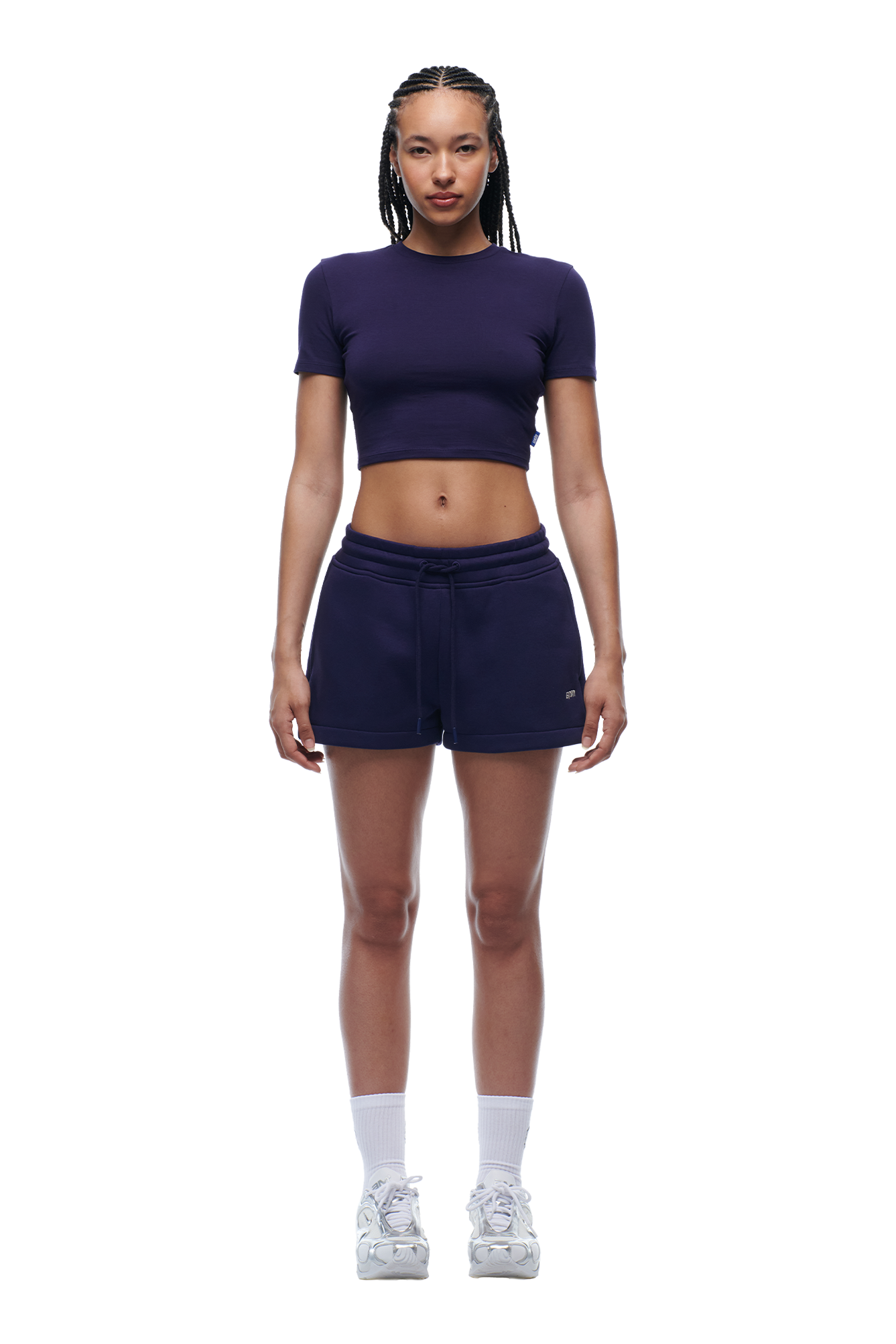 A woman with braided hair faces forward, wearing the 6PM WMNS CROPPED T-SHIRT ORBIT BLUE, navy athletic shorts, white socks, and sneakers against a white background.