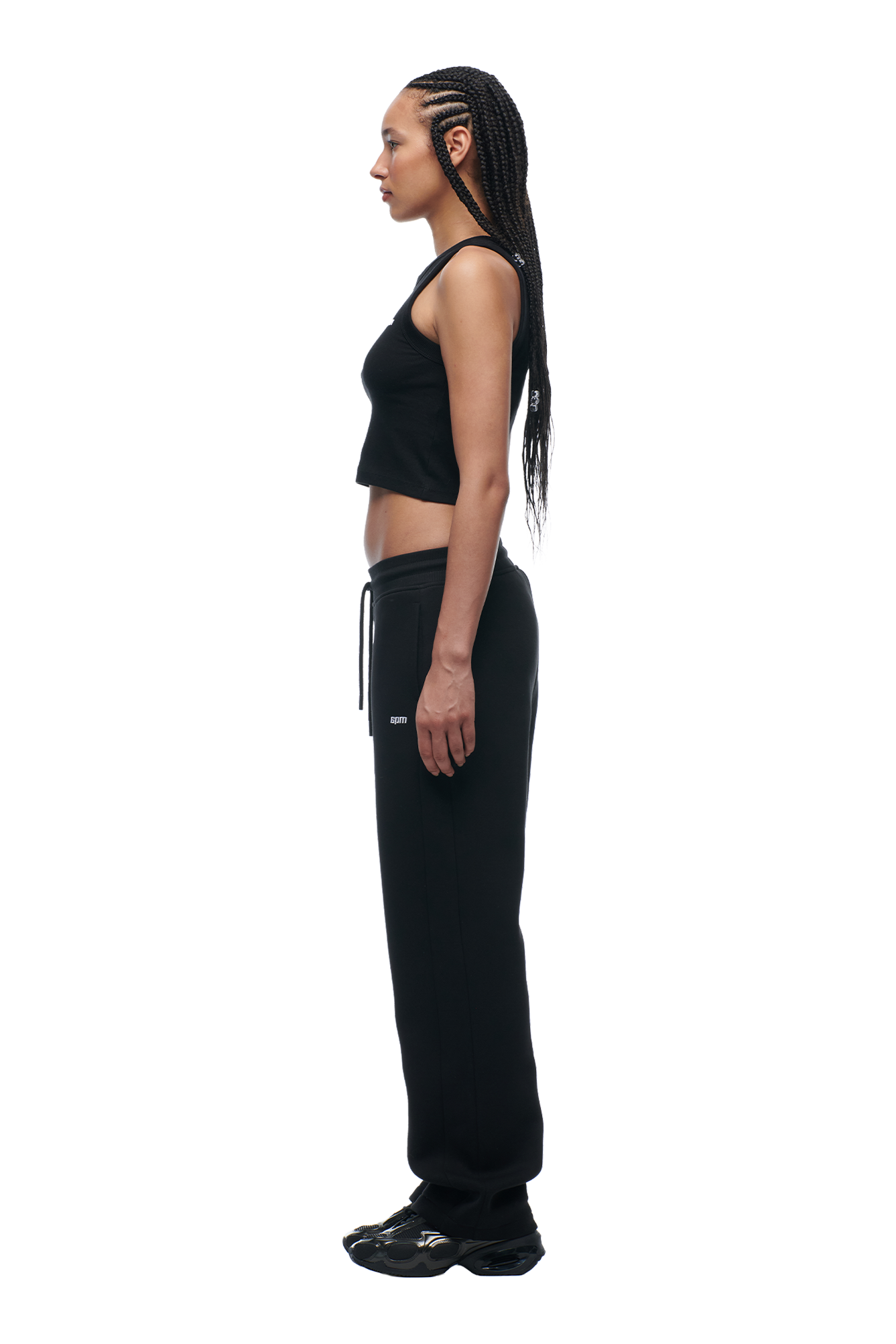 A woman with long braided hair stands in profile, wearing 6PMs WMNS OPEN LEG BLACK sweatpants and a black tank top with black sneakers, set against a white background.