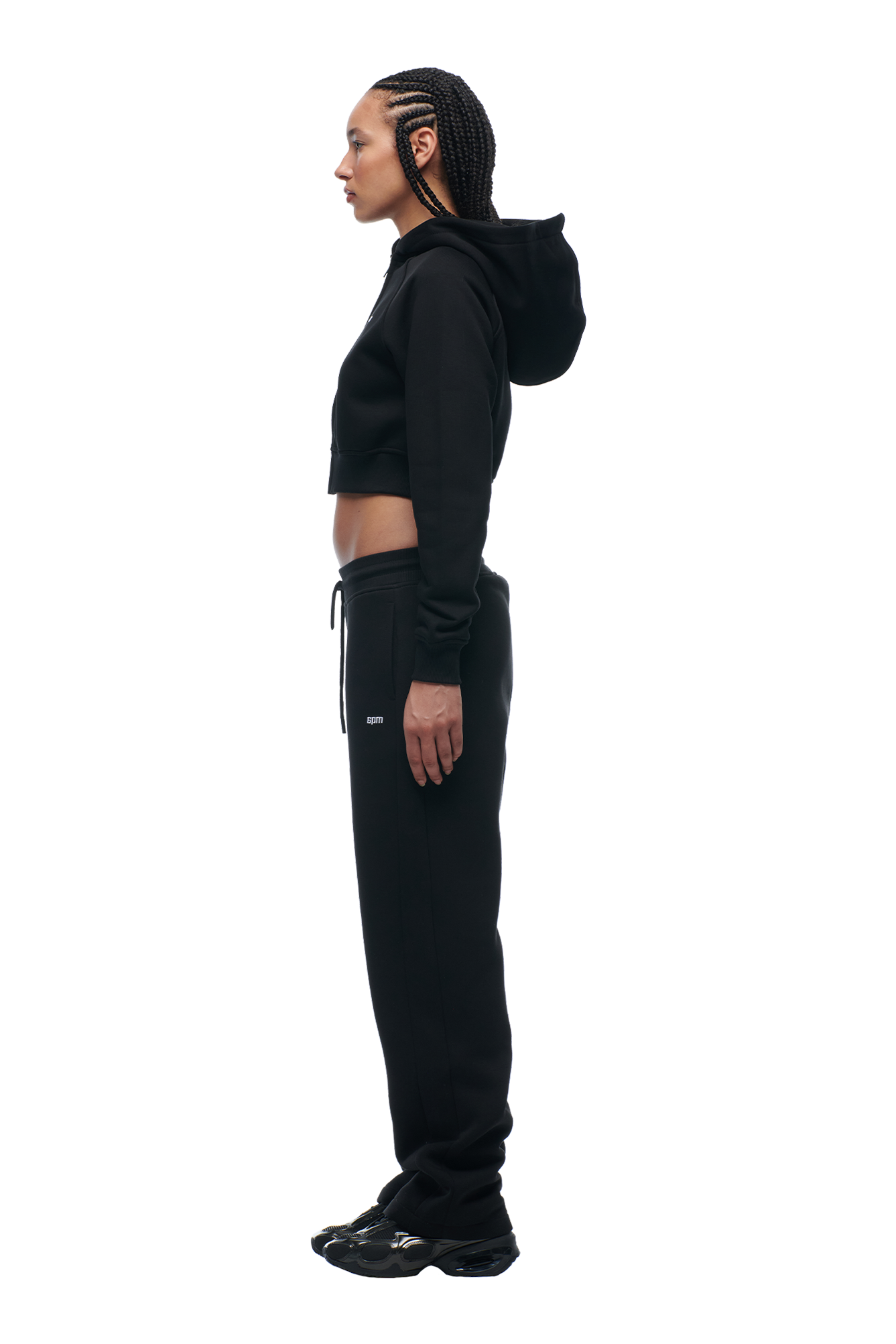 A person with braided hair stands in profile against a plain white background, wearing the 6PM WMNS Cropped Zip Hoodie Black, paired with black sweatpants and black shoes.