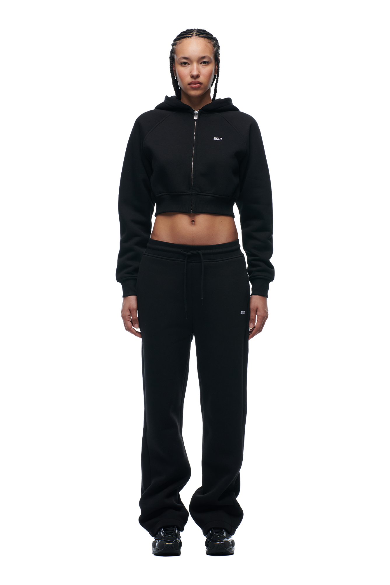 A woman with braided hair and a confident expression stands facing forward against a white background, wearing the 6PM WMNS CROPPED ZIP HOODIE BLACK, paired with black sweatpants and black sneakers.