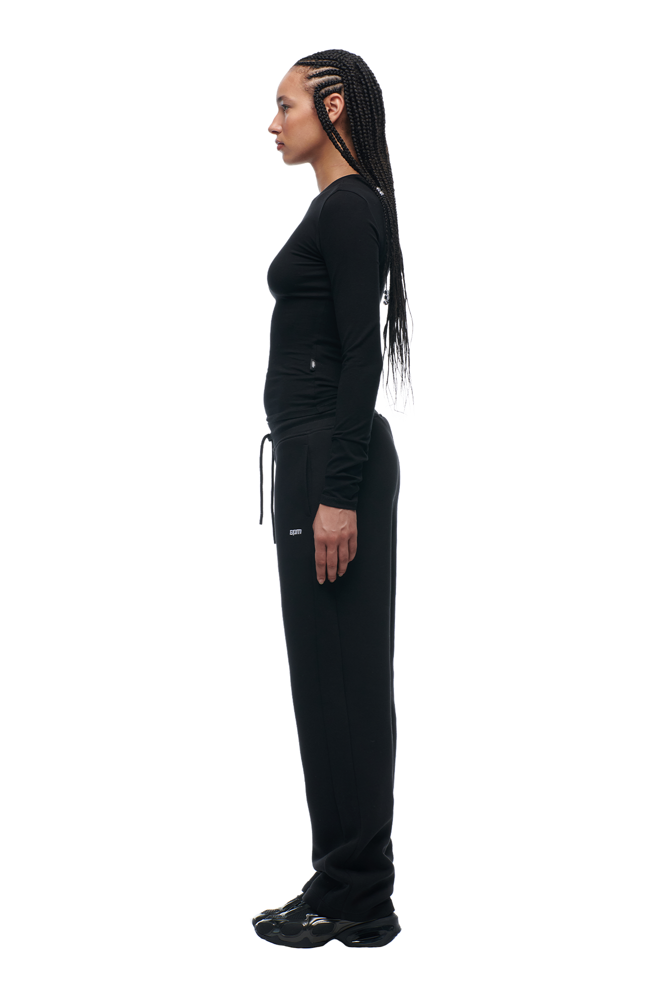 A woman with long braids stands in profile, facing left, wearing the 6PM WMNS LONGSLEEVE BLACK shirt with matching black sweatpants and shoes against a white background.