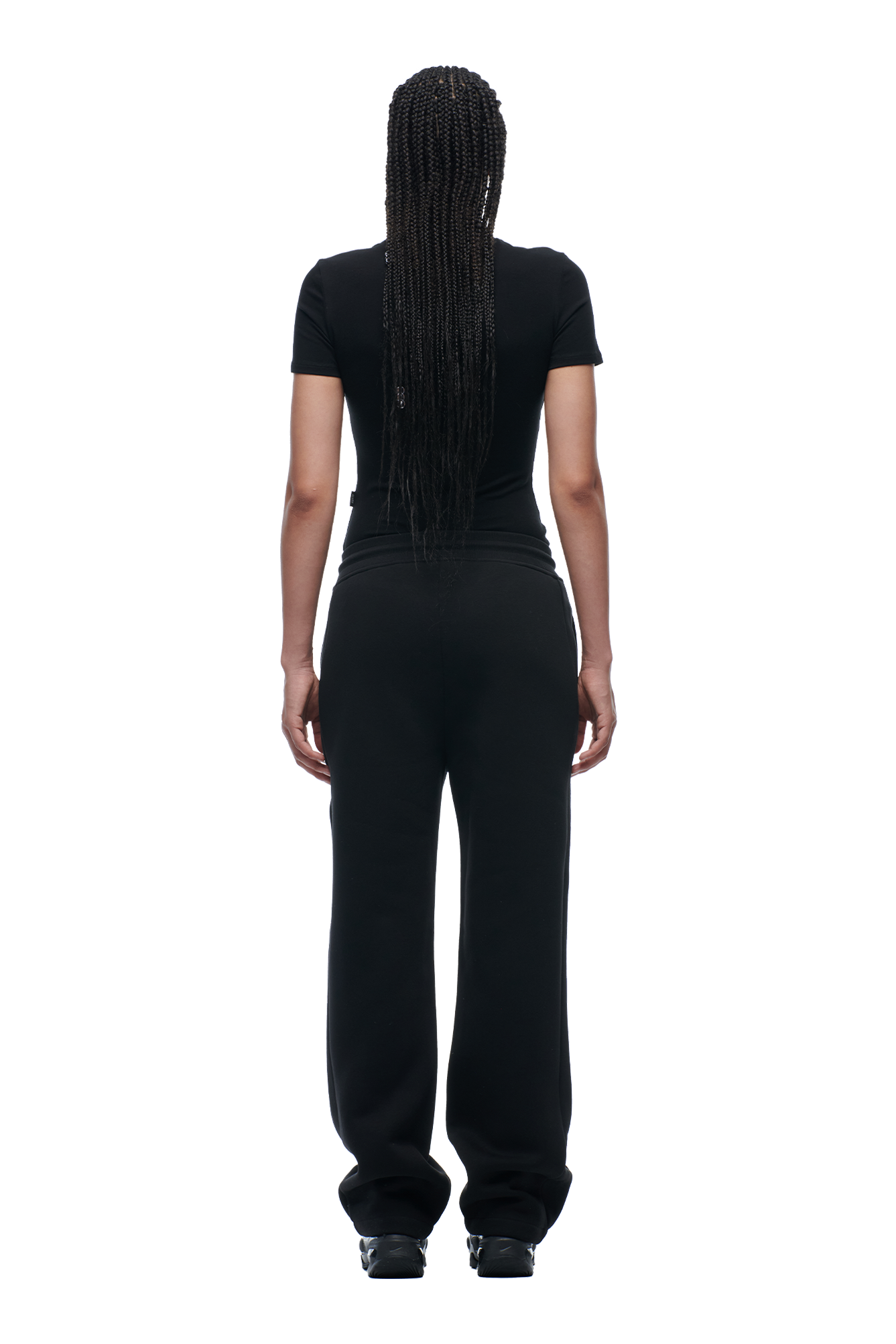 A person with long, braided hair faces away from the camera, wearing the 6PM WMNS T-SHIRT BLACK with loose black pants and black shoes against a white background.