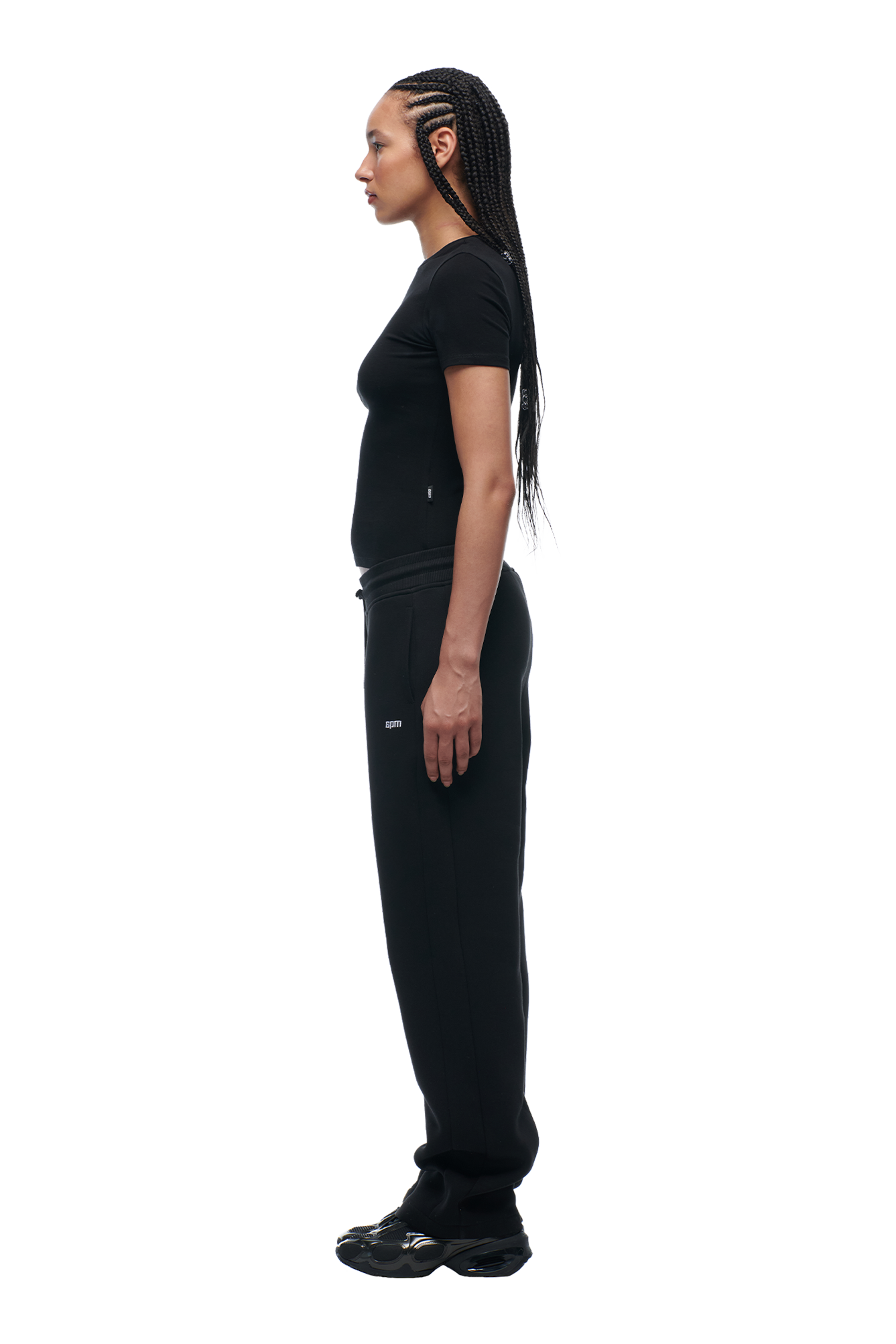 A woman with long braided hair stands in profile, facing left, wearing the 6PM WMNS T-SHIRT BLACK with matching black pants and shoes against a white background.