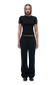 A woman with braided hair and a neutral expression wears the 6PM WMNS T-SHIRT BLACK, paired with black sweatpants and shoes. She stands facing forward against a plain white background.