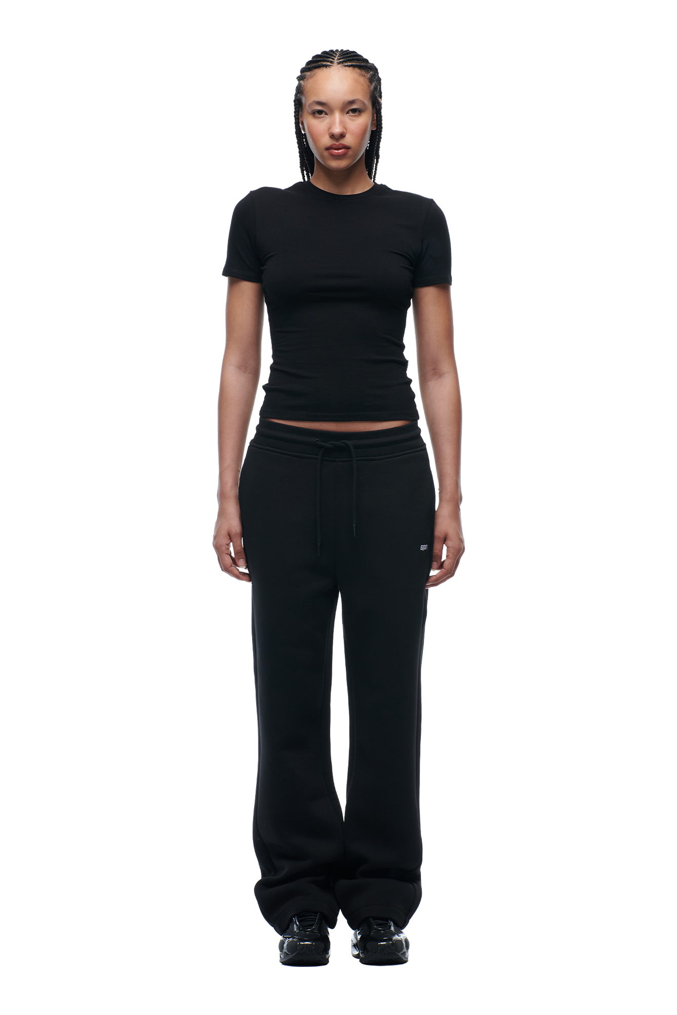 A woman with braided hair and a neutral expression wears the 6PM WMNS T-SHIRT BLACK, paired with black sweatpants and shoes. She stands facing forward against a plain white background.