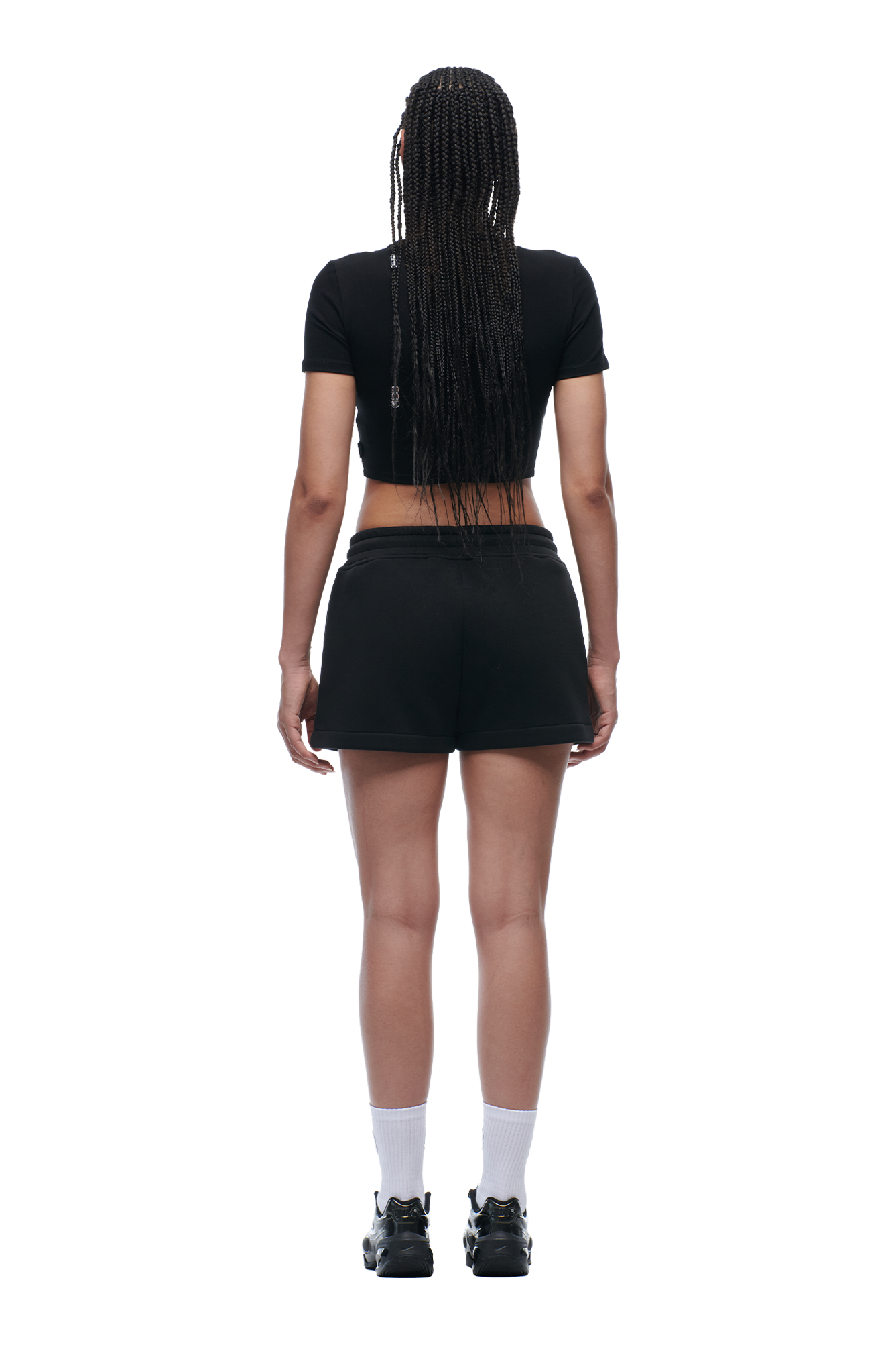 A woman with long braided hair wears the 6PM WMNS CROPPED T-SHIRT BLACK, black shorts, white socks, and black sneakers, standing away from the camera against a white background.