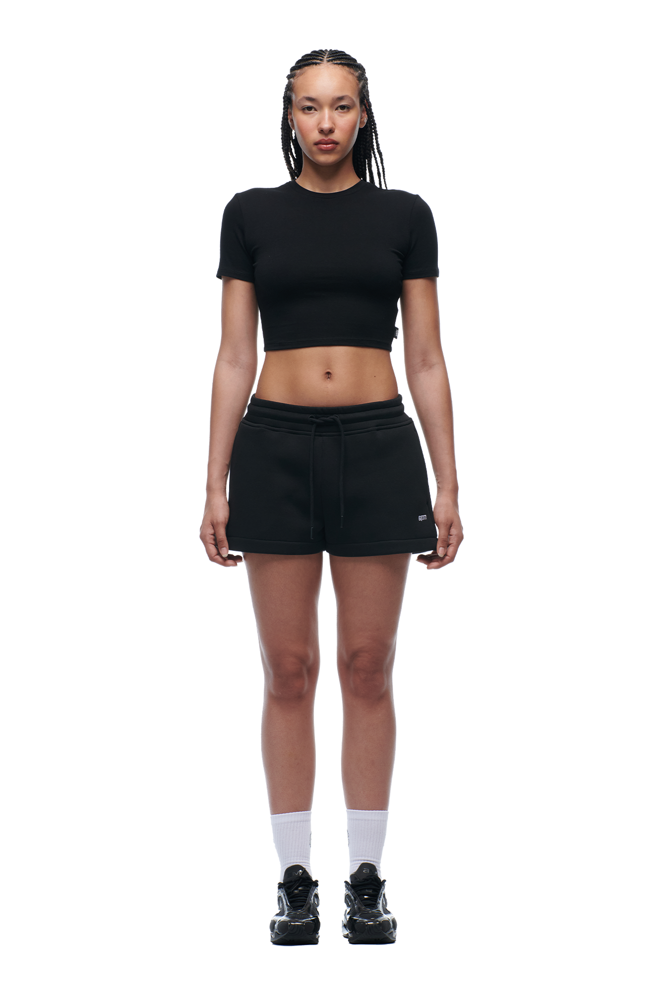 A woman with braided hair faces forward against a white background, wearing the 6PM WMNS CROPPED T-SHIRT BLACK, black shorts, white crew socks, and black sneakers.