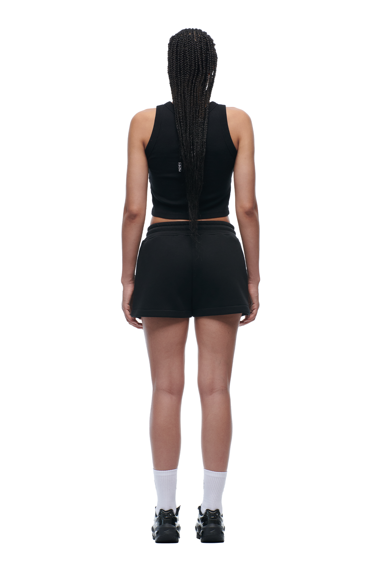 A person with long braided hair stands facing away, wearing a black sleeveless crop top, 6PM WMNS SHORTS BLACK, white crew socks, and black sneakers against a white background.