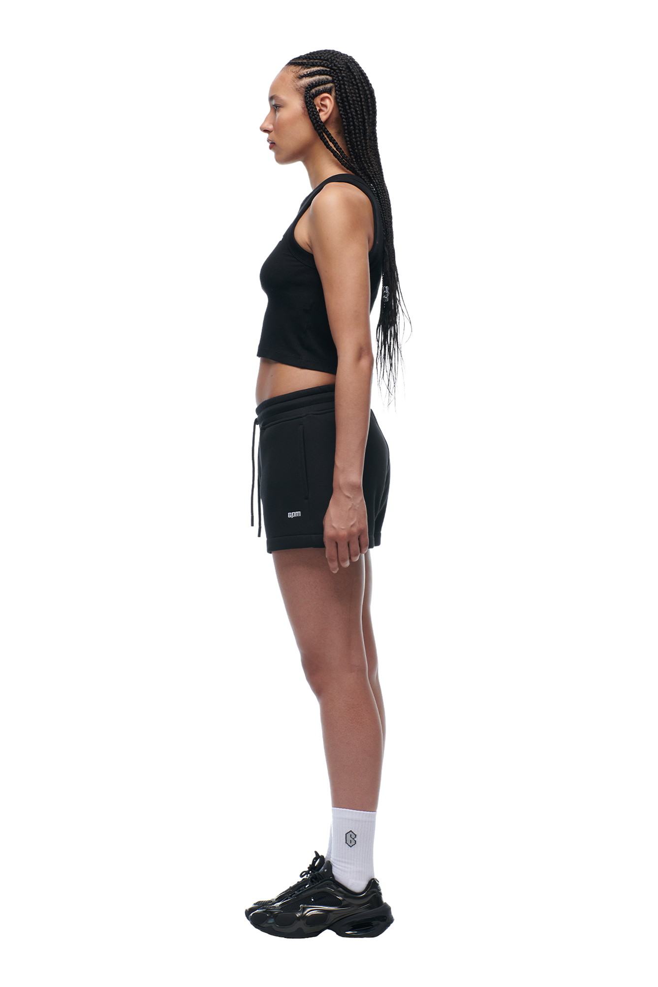 Woman in profile wears the 6PM WMNS SHORTS BLACK, paired with a black crop top, white socks, and black sneakers. She has long braided hair and stands against a white background.