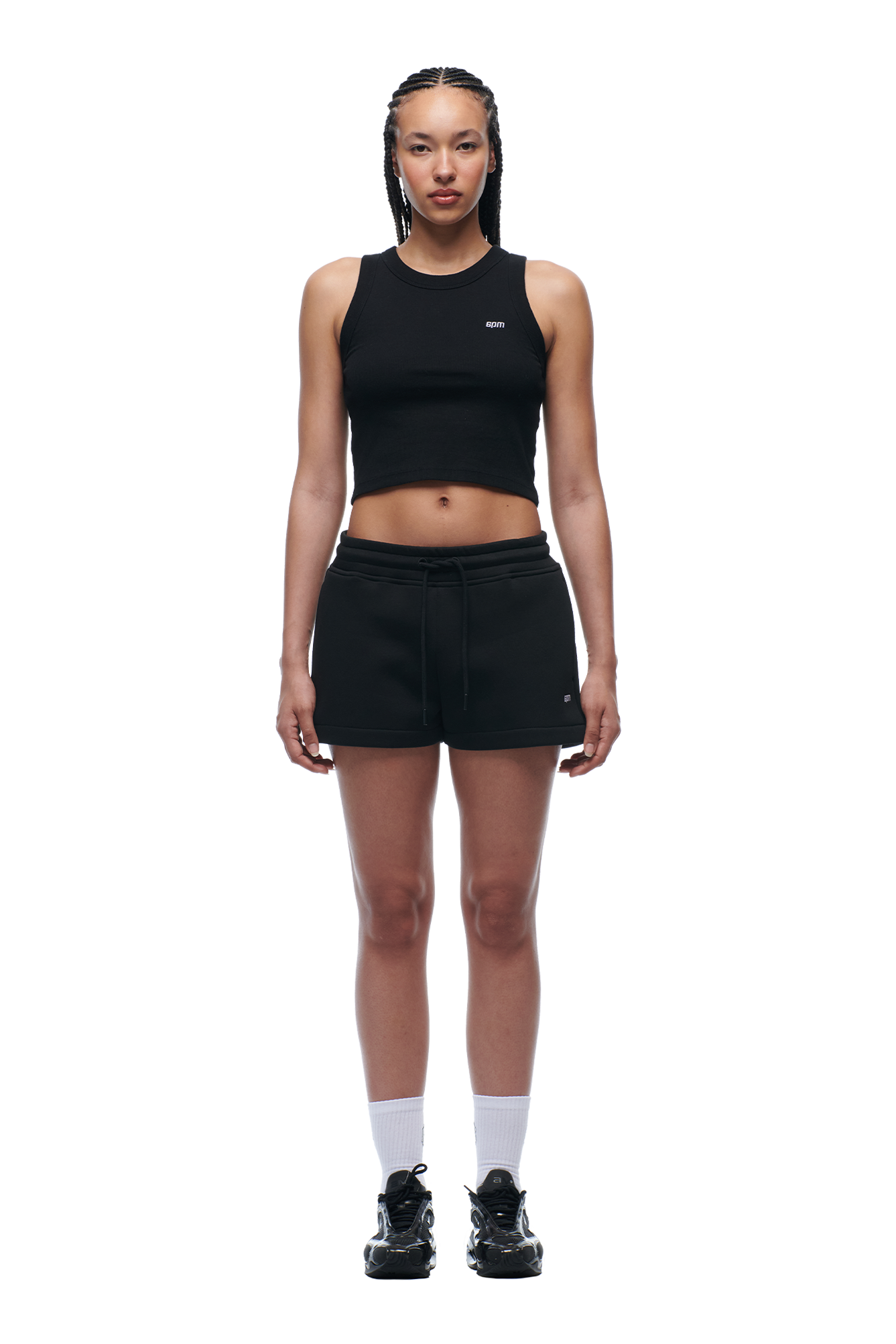 A woman with braided hair and a neutral expression faces forward against a plain white background, wearing 6PM’s WMNS SHORTS BLACK, a black sleeveless crop top, white socks, and black athletic shoes.
