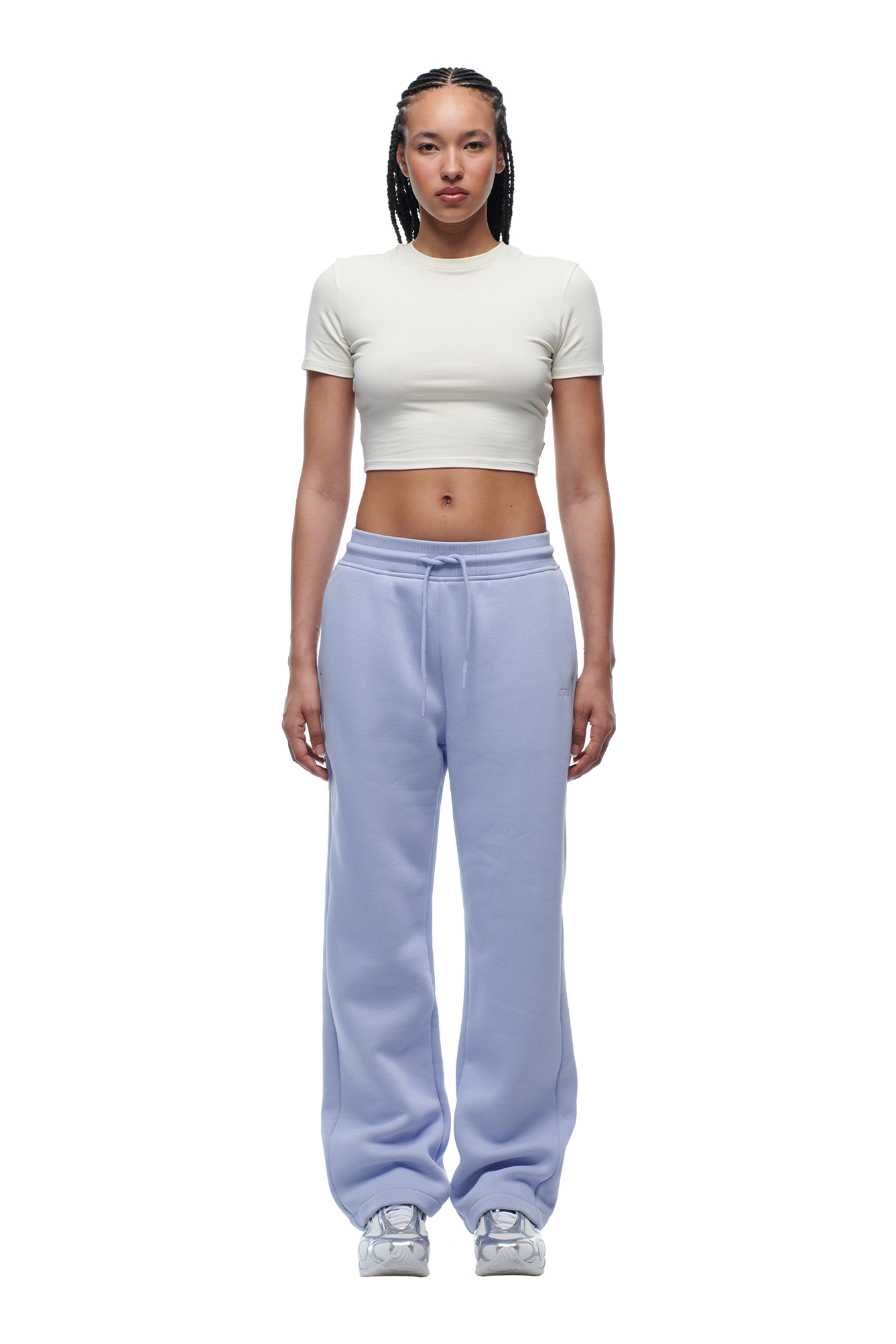 A woman with braided hair and a neutral expression stands against a white background, wearing the 6PM WMNS CROPPED T-SHIRT OFF-WHITE, paired with light blue sweatpants and white sneakers.