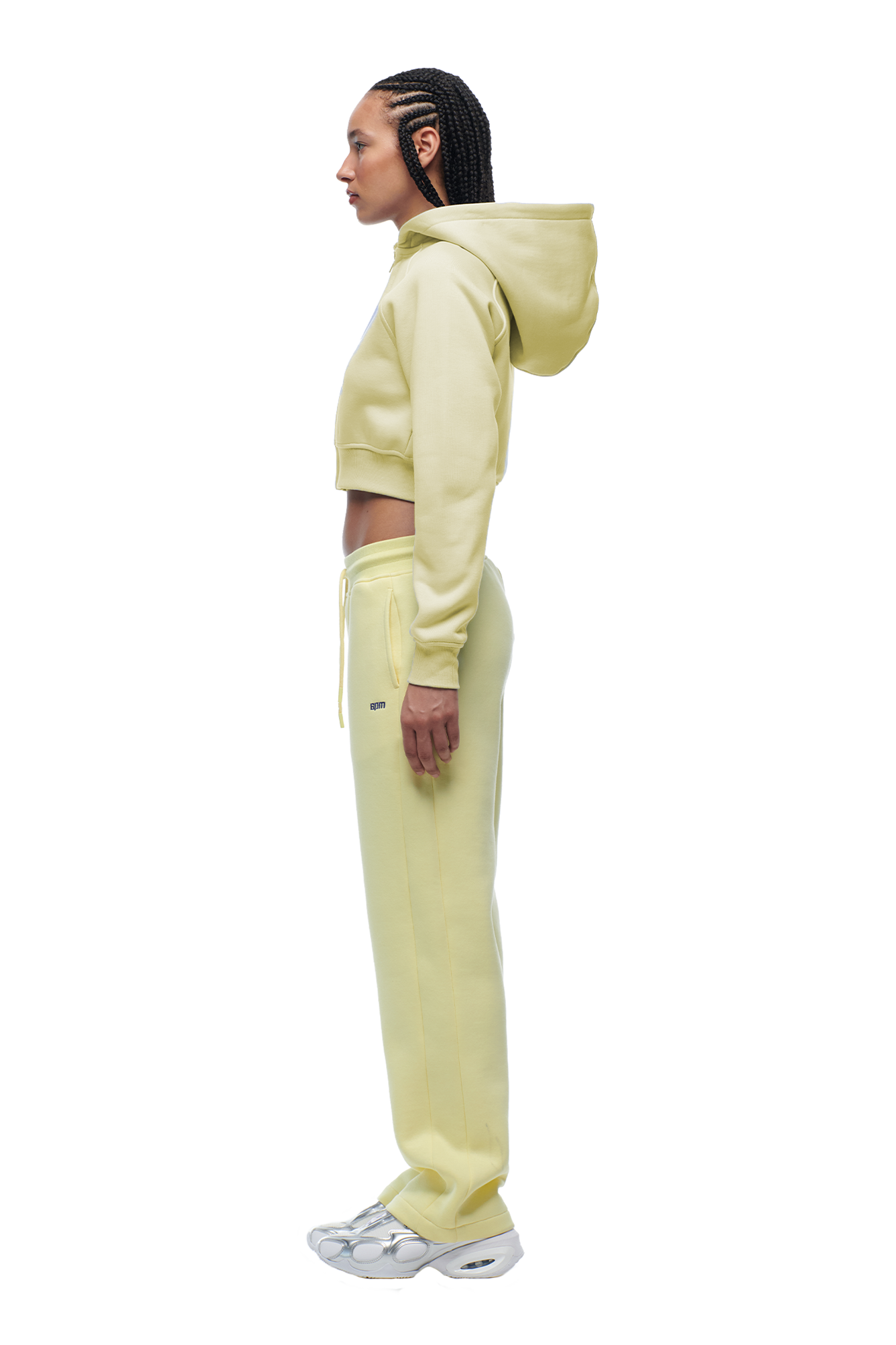 A woman with braided hair stands in profile, arms relaxed, wearing the 6PM WMNS CROPPED ZIP HOODIE in Buttercream with matching sweatpants and white sneakers. She faces left.