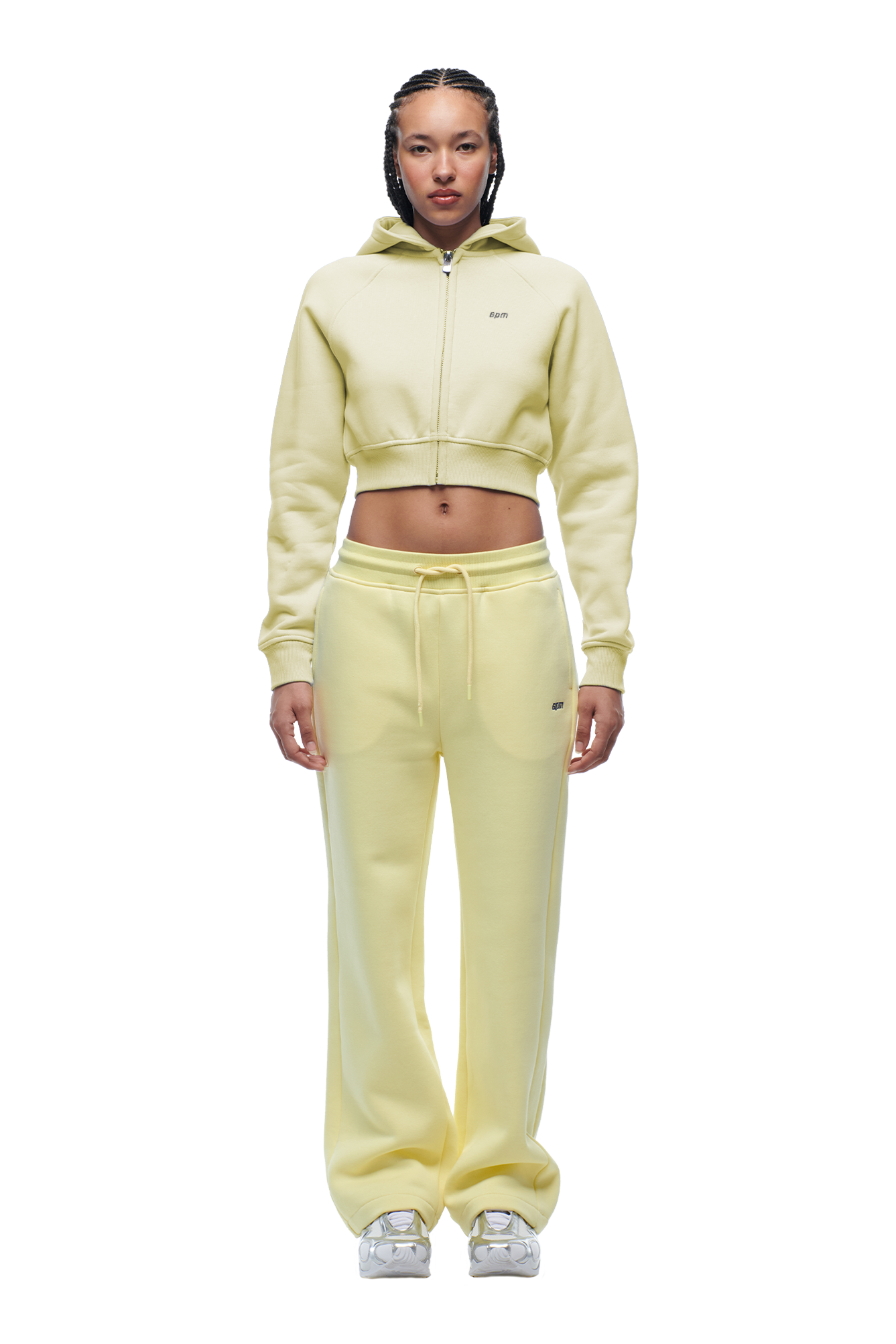 A woman with thin braids faces forward, wearing the 6PM WMNS Cropped Zip Hoodie Buttercream and matching sweatpants with white stripes, paired with white sneakers, looking directly at the camera.