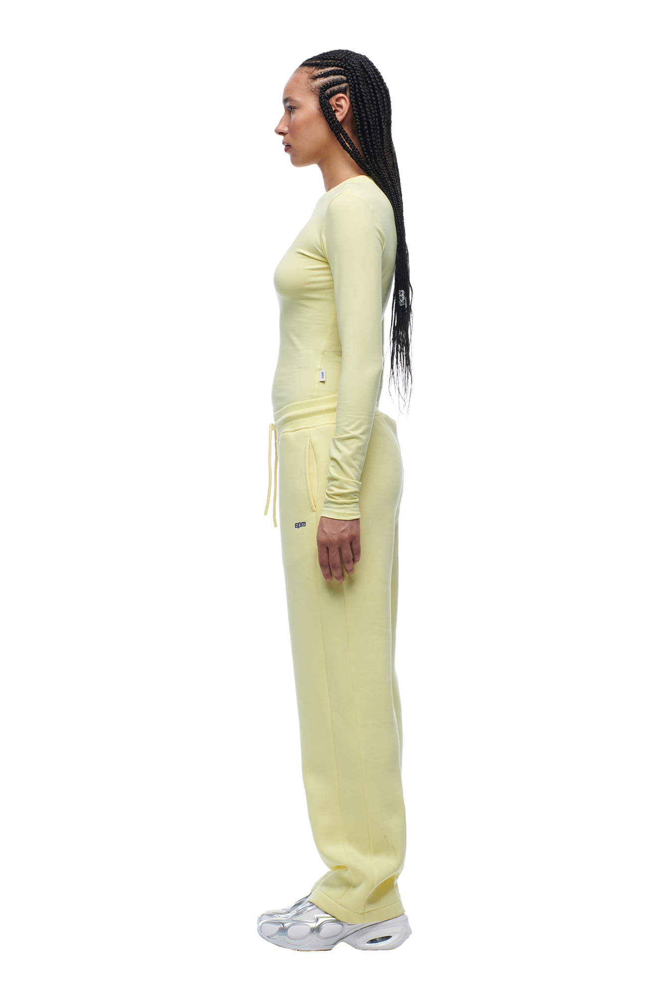 A woman with long braided hair stands in profile against a white background, wearing the 6PM WMNS LONGSLEEVE BUTTERCREAM top, matching yellow sweatpants with drawstrings, and white sneakers.