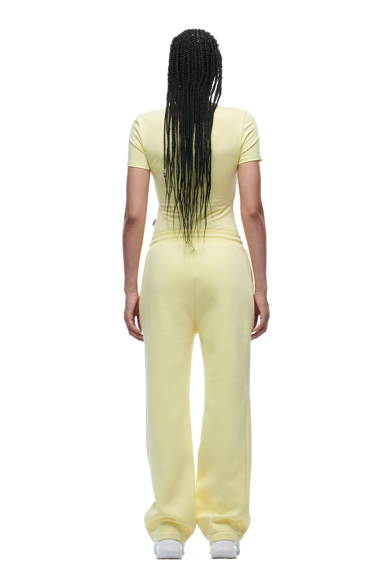 A person with long braided hair faces away, wearing the 6PM WMNS T-SHIRT BUTTERCREAM and matching loose pants with white sneakers against a white background.