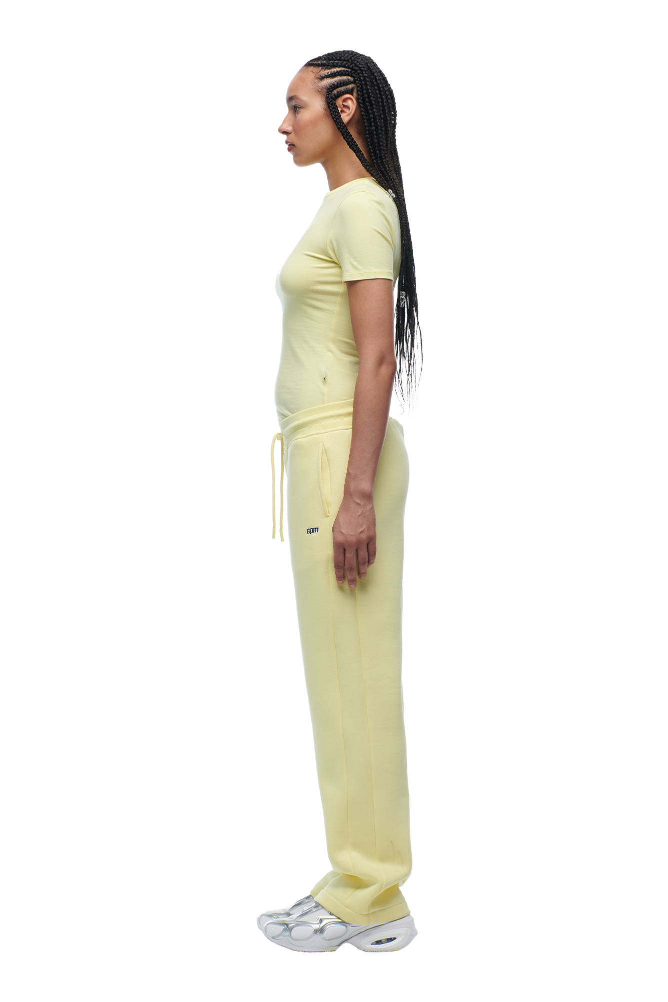 A person with long braided hair stands in profile, wearing the 6PM WMNS T-SHIRT BUTTERCREAM, matching sweatpants, and silver sneakers against a plain white background.
