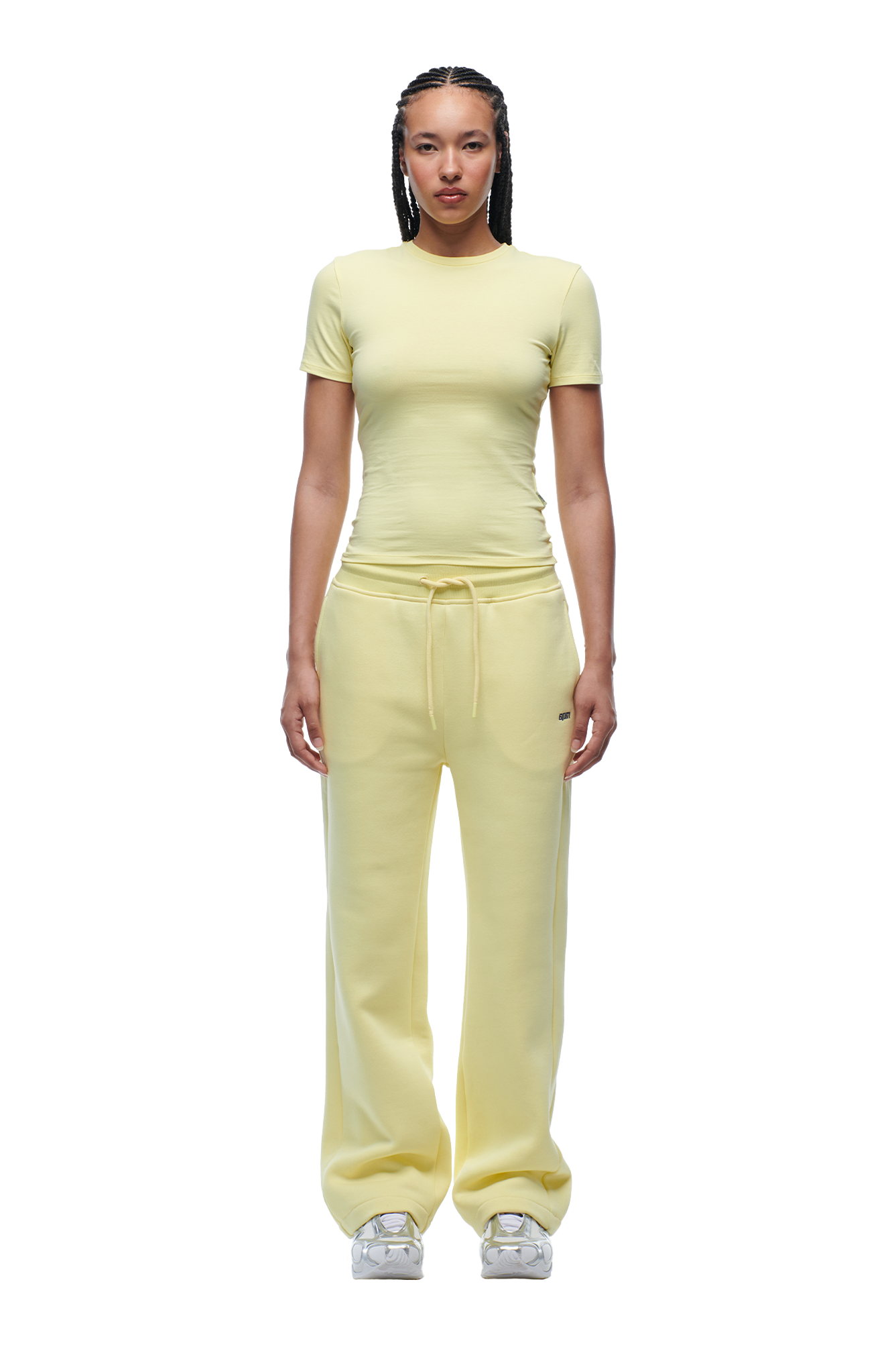 A person with braids and a neutral expression faces forward, wearing the 6PM WMNS T-SHIRT BUTTERCREAM and loose yellow sweatpants with a drawstring, paired with white sneakers.