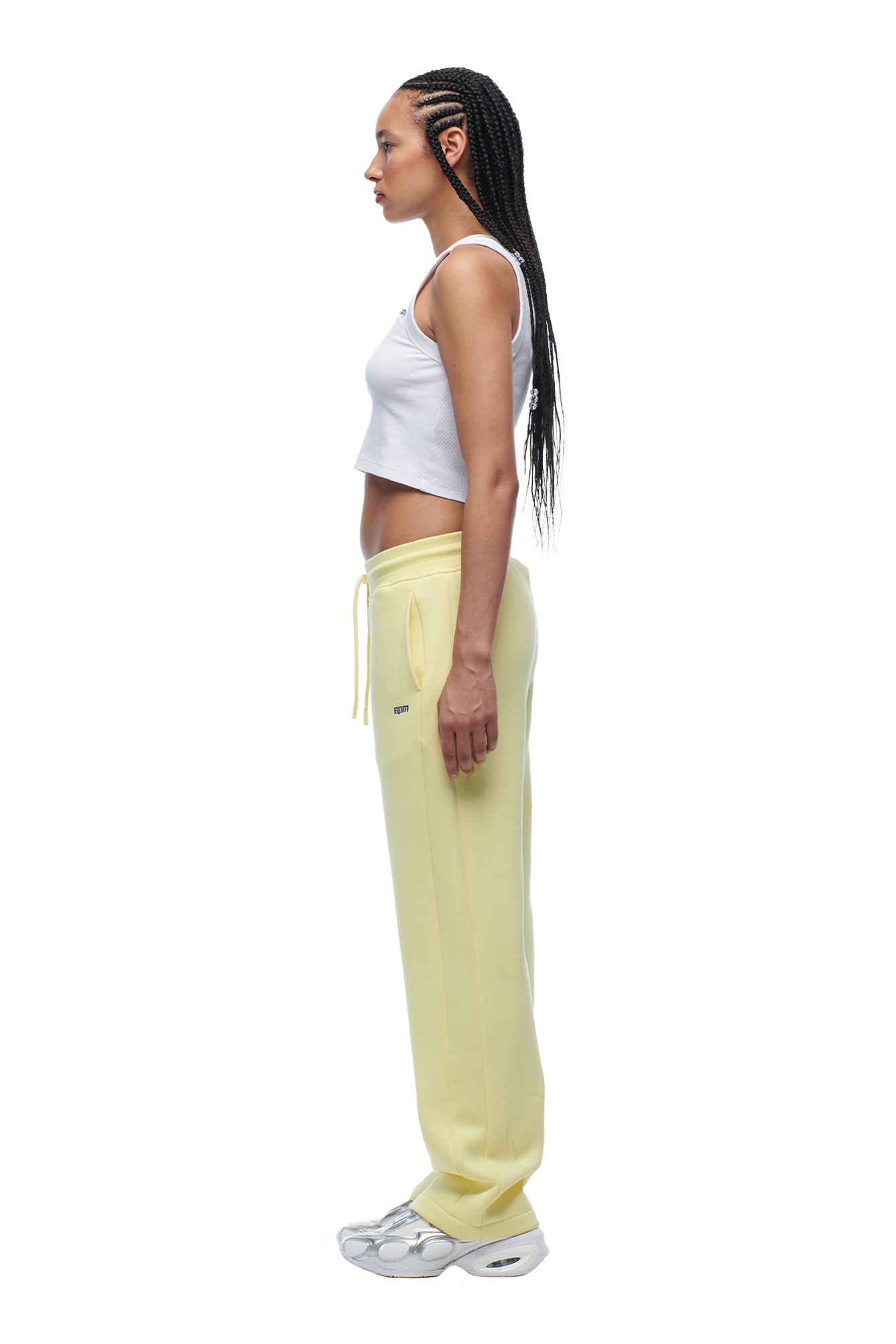 A woman with long braided hair stands in profile against a white background, wearing 6PM’s WMNS OPEN LEG BUTTERCREAM sweatpants paired with a white crop top and white sneakers.