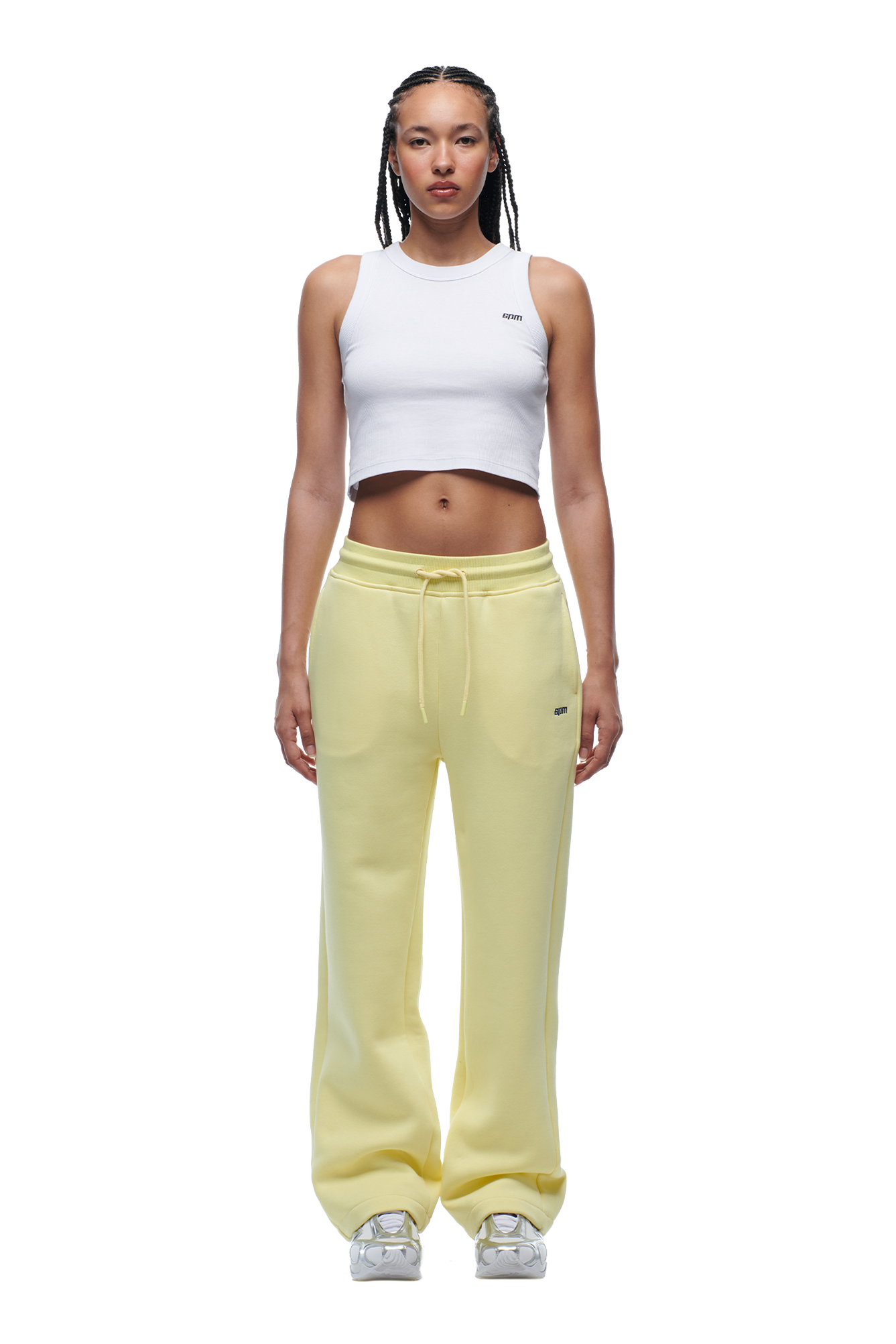 A woman with braided hair and a neutral expression faces forward, wearing the 6PM WMNS OPEN LEG BUTTERCREAM sweatpants, a white cropped tank top, and white sneakers.