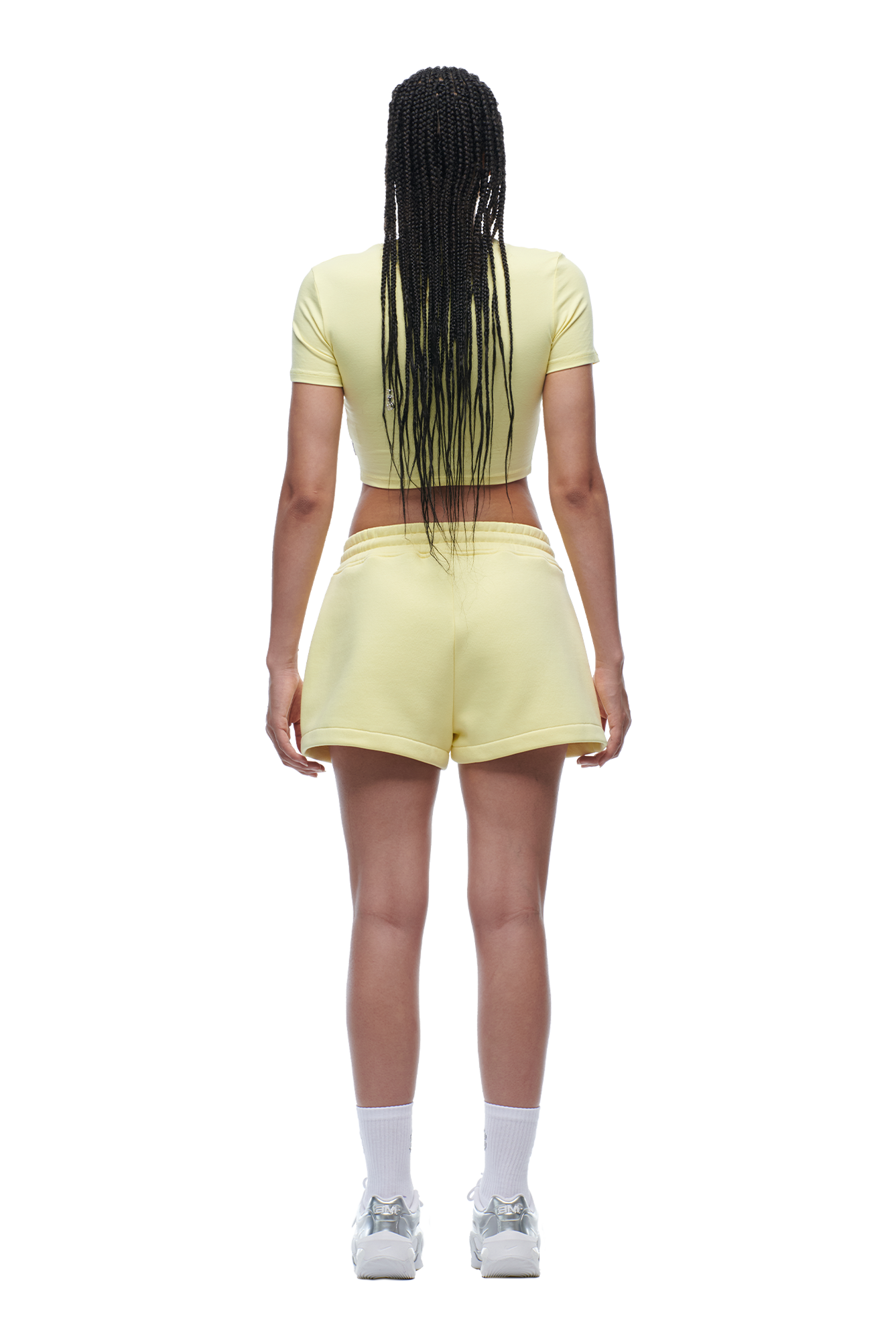 A person with long braided hair faces away, wearing the 6PM WMNS CROPPED T-SHIRT in Buttercream, paired with matching shorts featuring side stripes, plus white socks and sneakers.