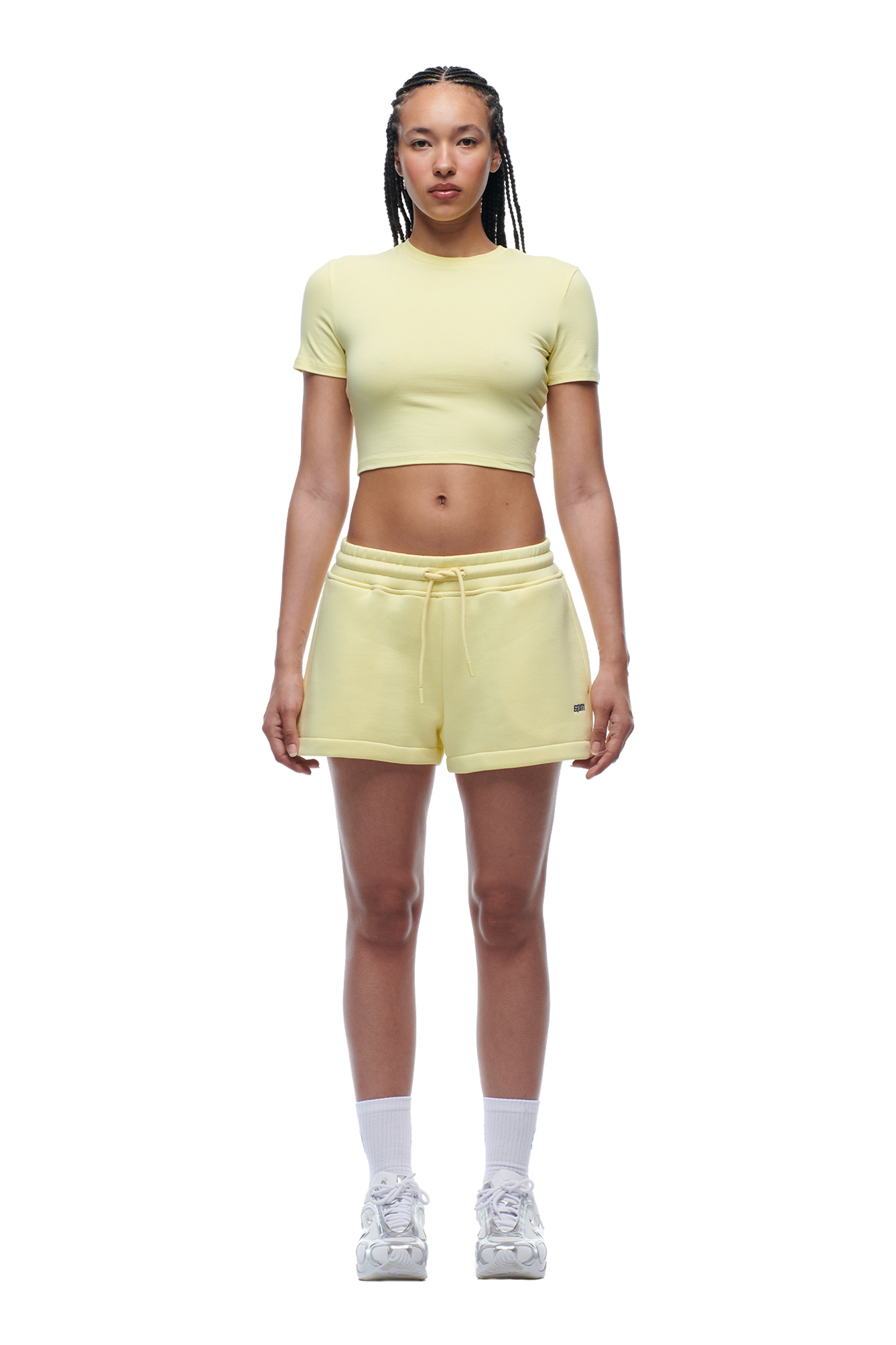 A person with braided hair and a neutral expression faces forward, wearing the 6PM WMNS Cropped T-Shirt Buttercream, matching yellow drawstring shorts, white socks, and white sneakers.