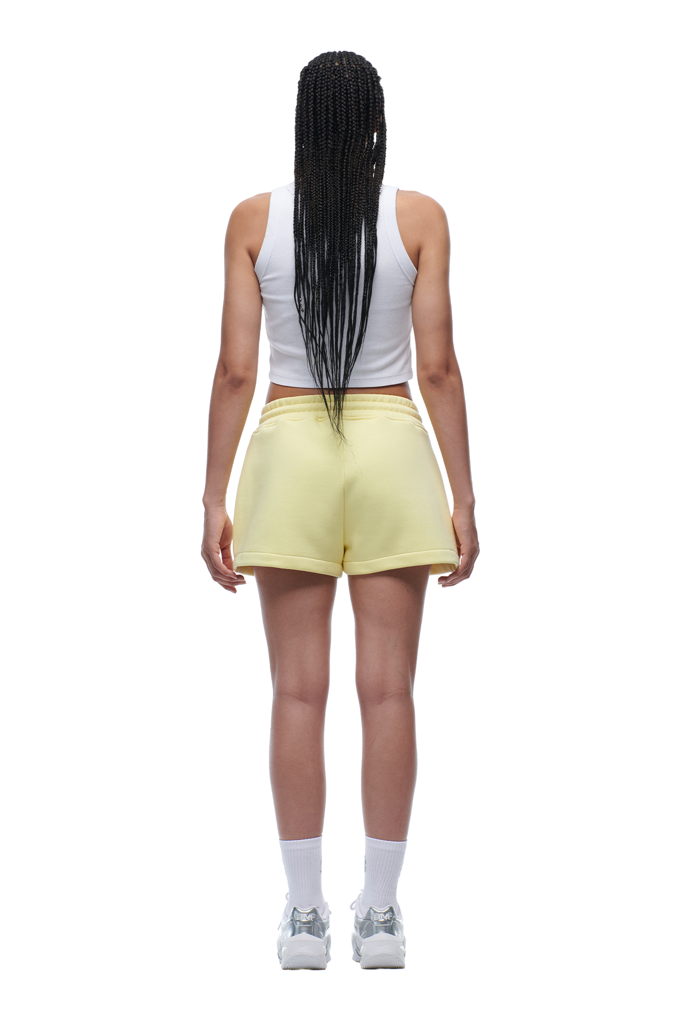 A person stands facing away from the camera against a white background, wearing 6PM WMNS SHORTS BUTTERCREAM, a white tank top, white socks, and sneakers.