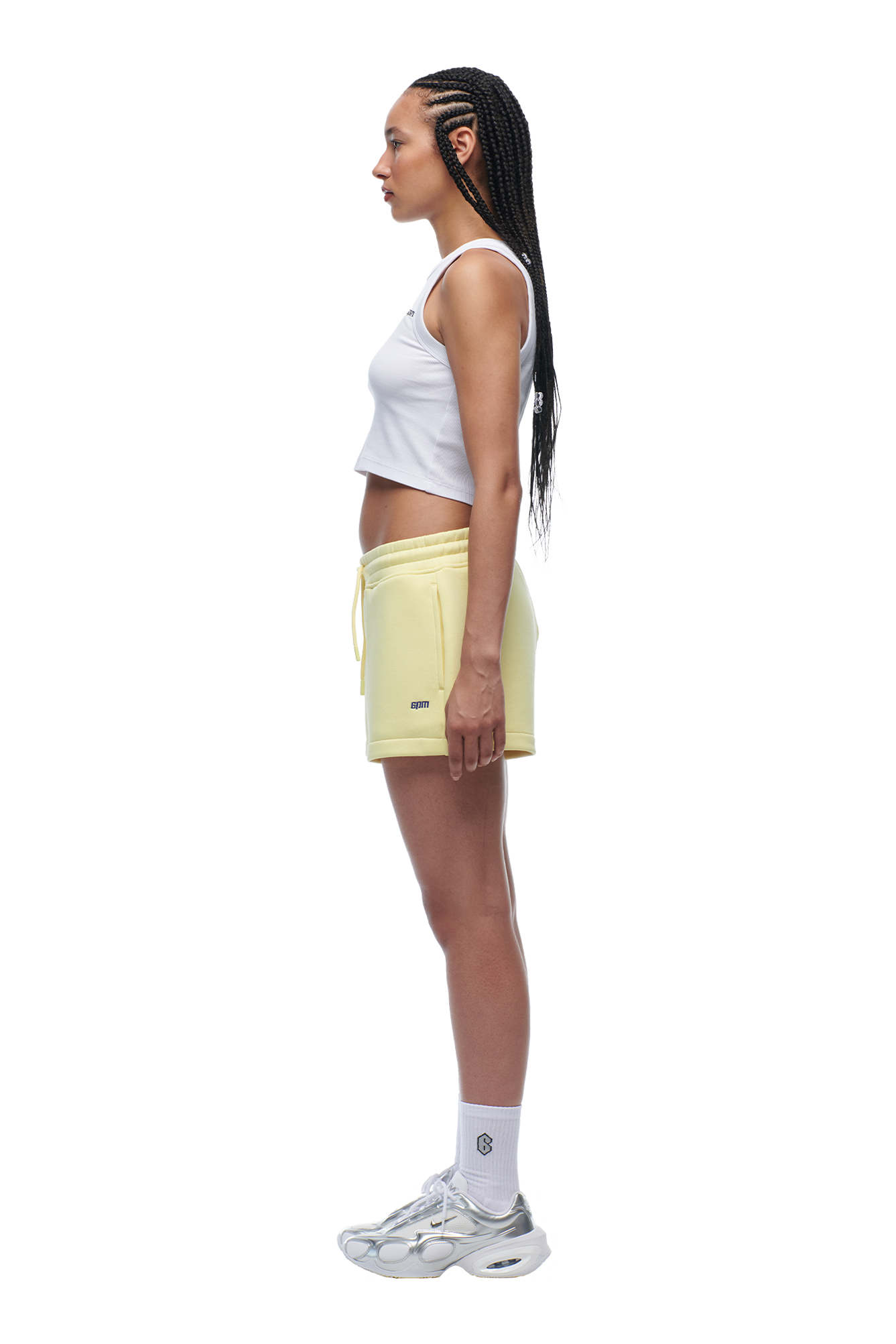 A woman with long braided hair stands in profile, wearing a white crop top, 6PM WMNS SHORTS BUTTERCREAM, white crew socks, and white sneakers.